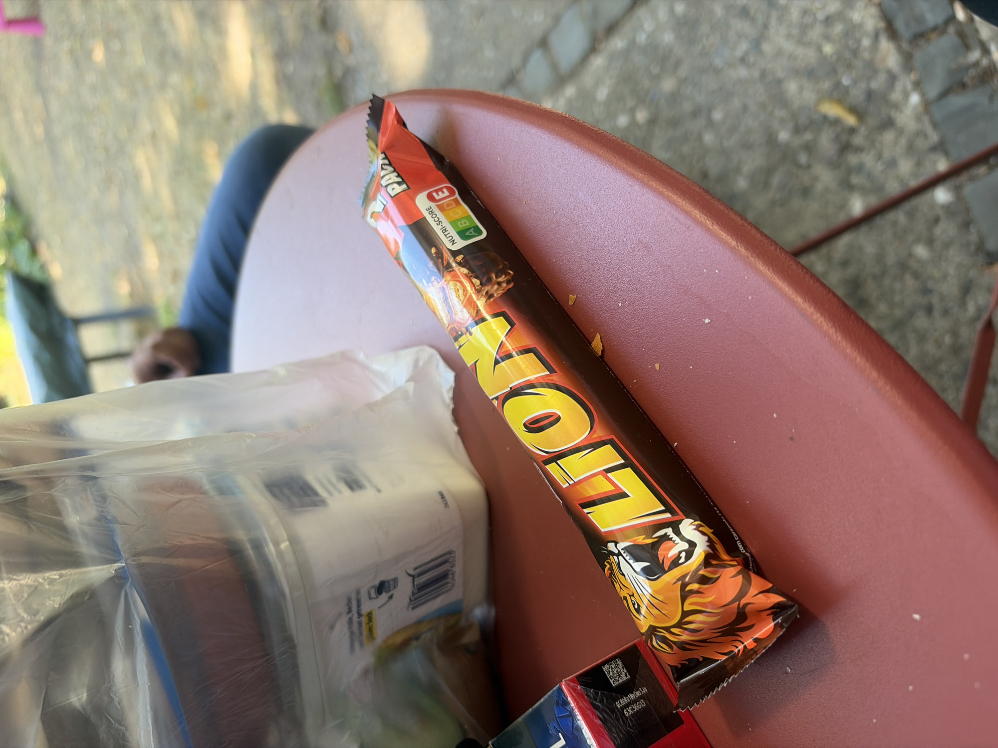 A partially obscured plastic bag and a Lion chocolate bar are positioned on a red, circular table outdoors. The chocolate bar is wrapped in a dark brown and orange wrapper featuring a roaring tiger's head and the "LION" logo in large, yellow, distressed lettering. A Nutri-score rating of "C" is visible on the wrapper. In the background, a person's legs are visible, clad in blue jeans, and they appear to be sitting. The ground is a mix of gravel and paving stones, suggesting an outdoor setting like a park or beer garden. The lighting indicates it is daytime. The image was taken in Dusseldorf, Germany.