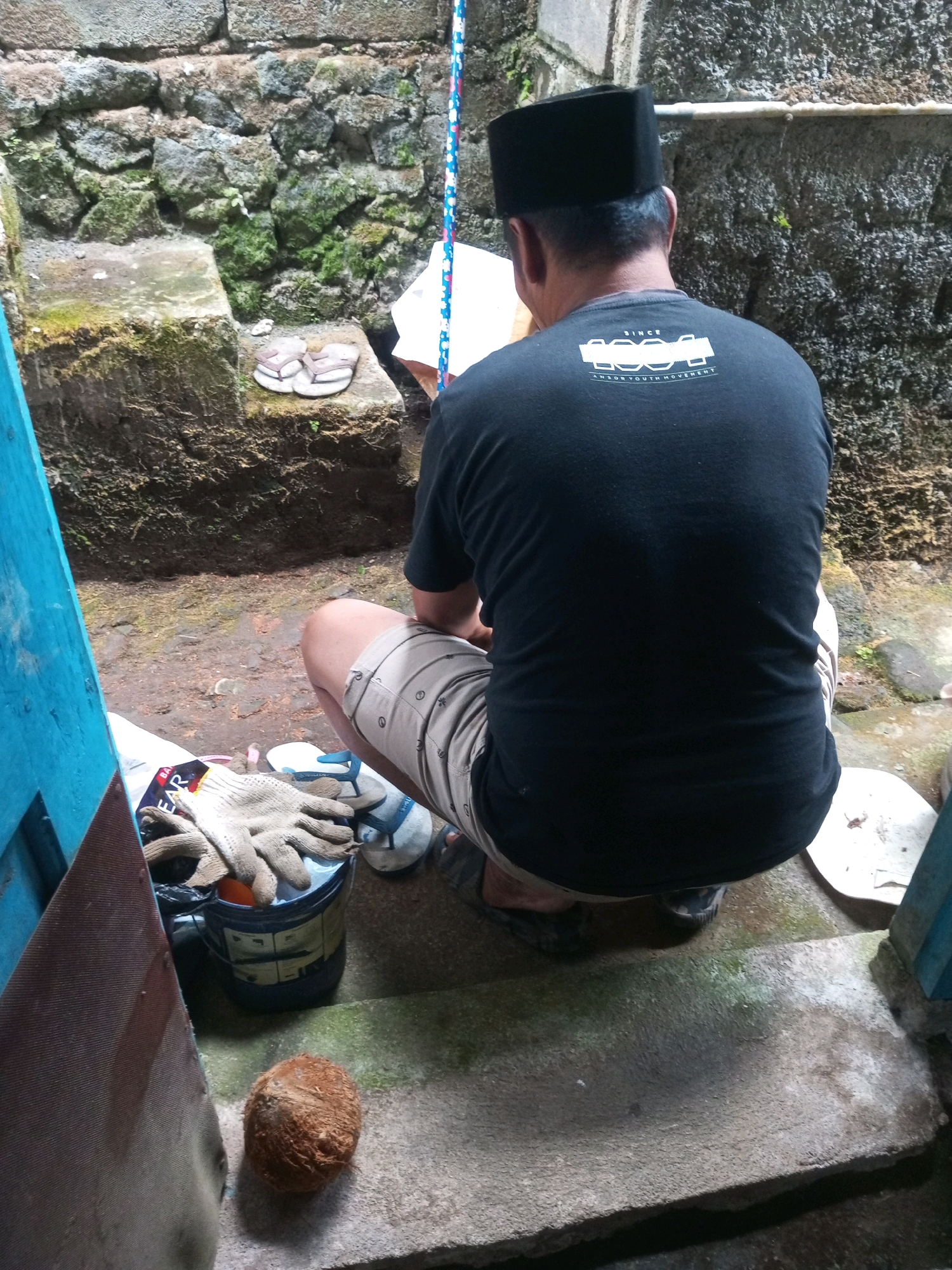 A man wearing a black peci and a black t-shirt with a white graphic on the back is squatting on a concrete step. He is also wearing light-colored shorts with a pattern. In front of him, on the ground, is a whole coconut. To his left is a blue bucket with a pair of work gloves and a plastic bag on top of it. Behind him is a textured stone wall. Further behind the man and to the left, there is a stone staircase with a pair of flip-flops resting on it. The environment appears to be an outdoor space with dirt and moss on the ground and the walls. The scene is lit by natural light.