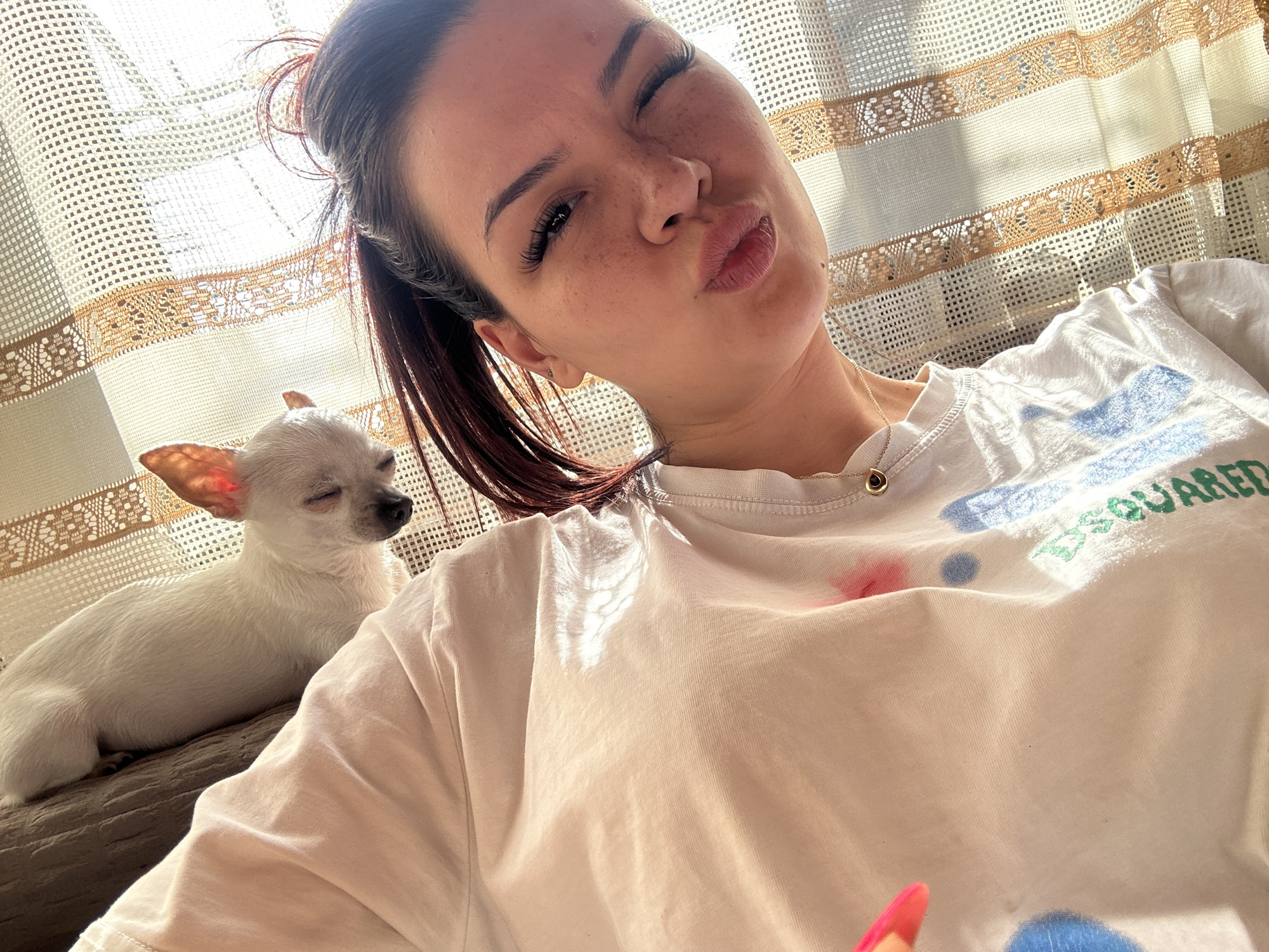 A woman poses for a selfie indoors with a small, white chihuahua. The woman is light-skinned with brown hair in a high ponytail and is wearing a white t-shirt with colorful spots on it. She is winking and pouting. The chihuahua, located to the left of the woman, is lying down with its eyes closed. The background includes a patterned curtain with sunlight streaming through it. The location is not discernable from the image.