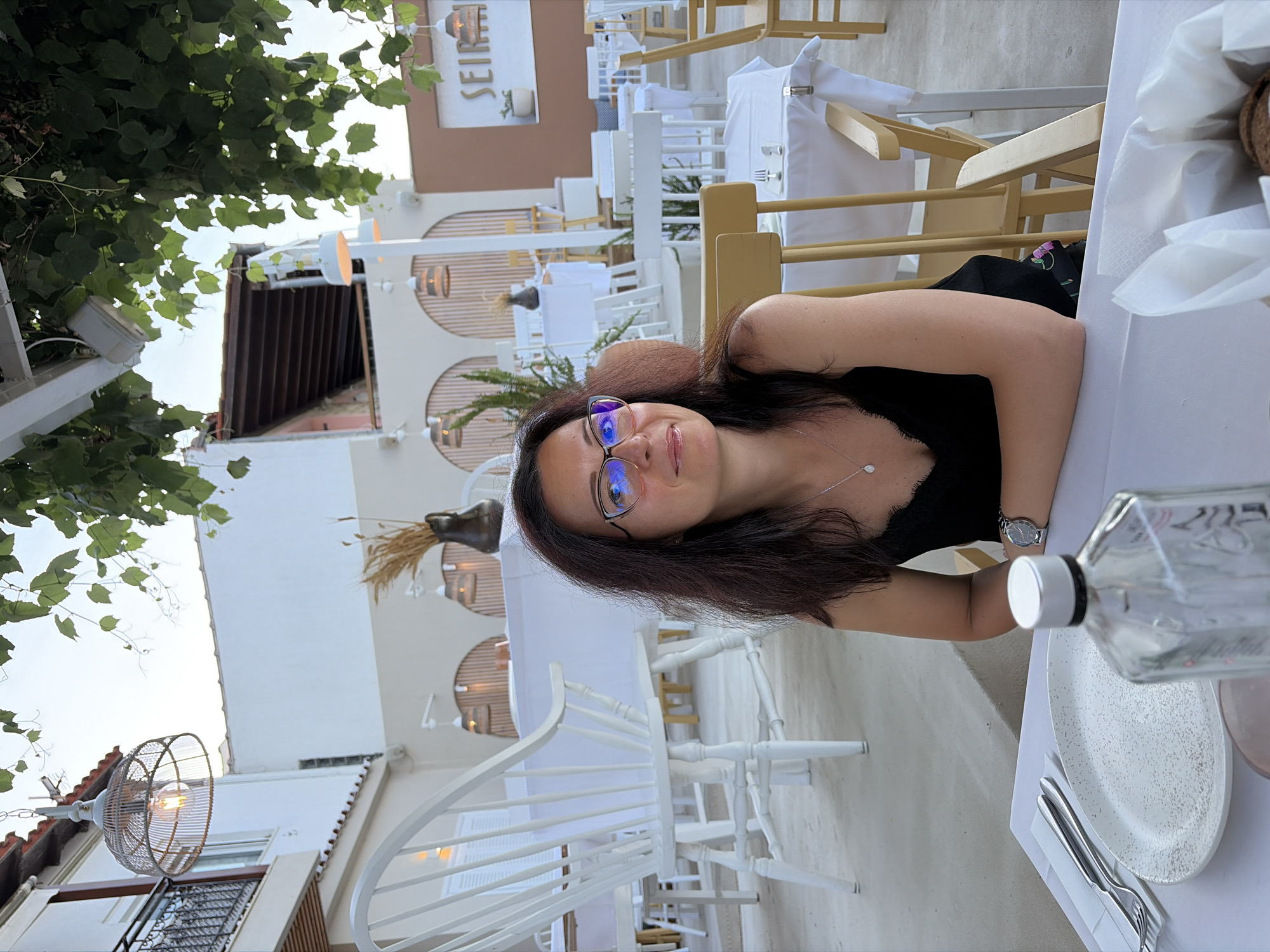 This image depicts a woman seated at a table in an outdoor restaurant. She is wearing glasses and a black top, and has dark hair. A water bottle and a white plate with silverware are visible on the table in front of her. The restaurant setting is airy and appears to be during the daytime, with white furniture and a bright sky. The scene suggests a casual dining experience. The word "SEIR" is visible on a sign in the background.