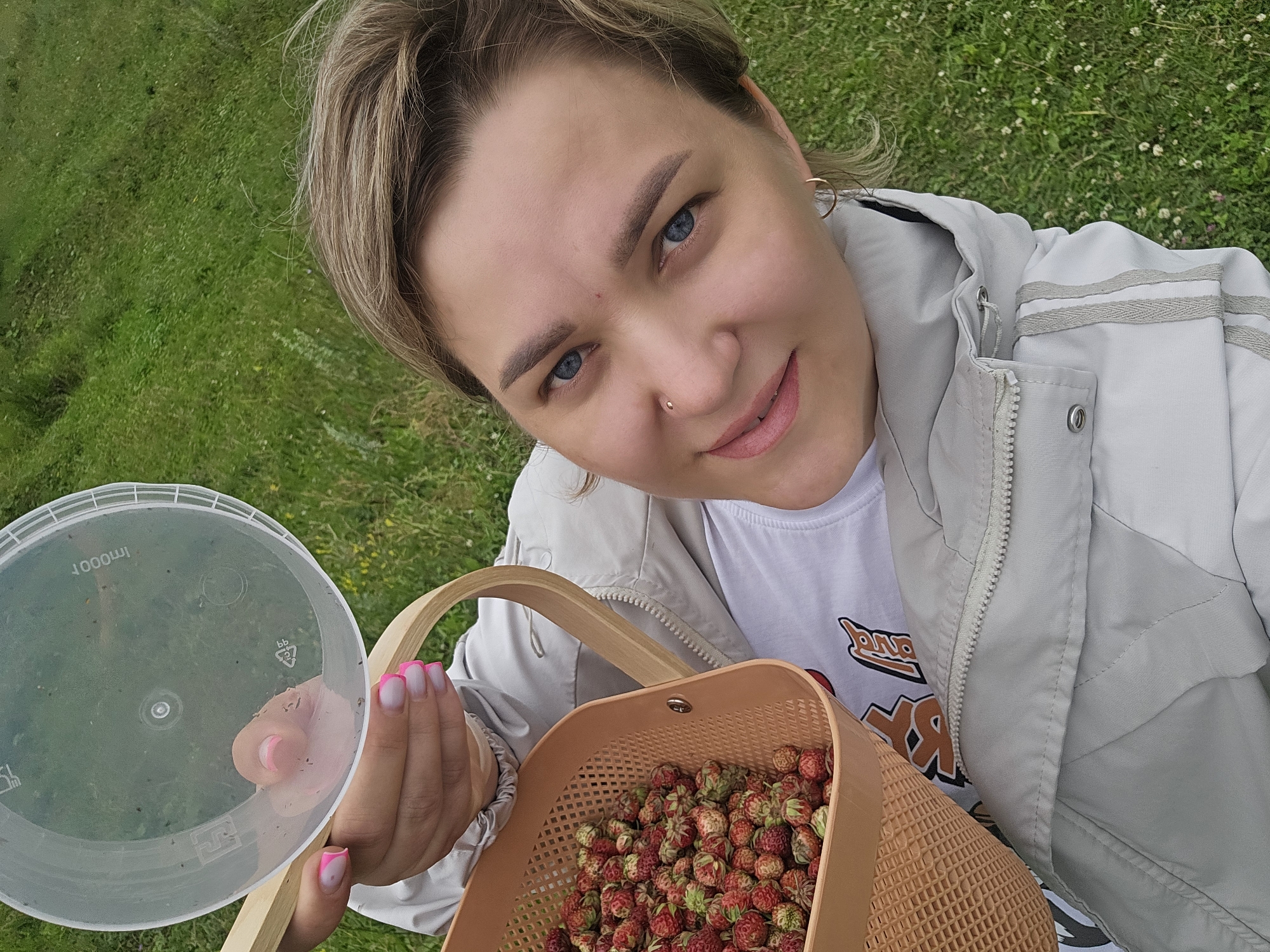 A woman is taking a selfie outdoors, holding a transparent lid in her left hand and a basket full of strawberries in her right. She is wearing a light-colored jacket over a white t-shirt. Her hair is short and blonde, and she has blue eyes and light makeup. The basket, made of light brown plastic, is brimming with small, red and green strawberries. The lid she holds appears to be from a container, with some smudges on its surface.
The background consists of lush green grass, indicating an outdoor setting. The lighting suggests it is daytime, possibly under an overcast sky or in shade, given the soft illumination. The woman's expression is a bright, engaging smile, and her blue eyes are looking directly at the camera. Her nails are painted a vibrant pink with a white French tip.
The scene evokes a sense of leisure and outdoor activity, likely berry picking. The overall mood is cheerful and relaxed. The visible text on the lid is "1000ml", and a recycling symbol with the number "5" is also visible. The location context is provided as Kalinovka, Russia.