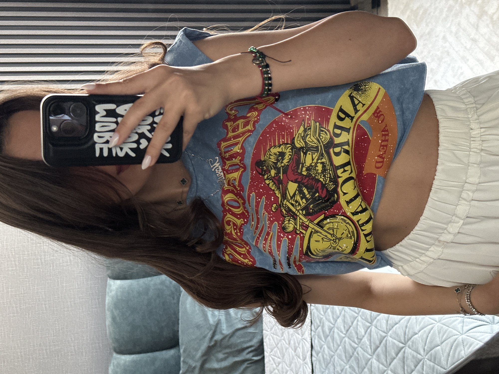 A young woman with long, dark brown hair is taking a mirror selfie in what appears to be a bedroom in Sofia, Bulgaria. She is wearing a light blue cropped t-shirt with a graphic print of a motorcycle rider. The graphic is colorful, featuring a red and yellow design with flames. She is also wearing white, high-waisted pants. On her left wrist, she has two bracelets, one with a clover charm. On her right wrist, she has a beaded bracelet with green and black beads. She is holding a black iPhone in her right hand, with the screen facing the mirror, displaying the words "MORE WORK LESS WORRY". The background includes a bed with light blue quilted bedding, a headboard, and a textured light grey wall. Vertical striped wallpaper or blinds are visible on the right side of the frame. The overall lighting suggests an indoor, daytime setting.