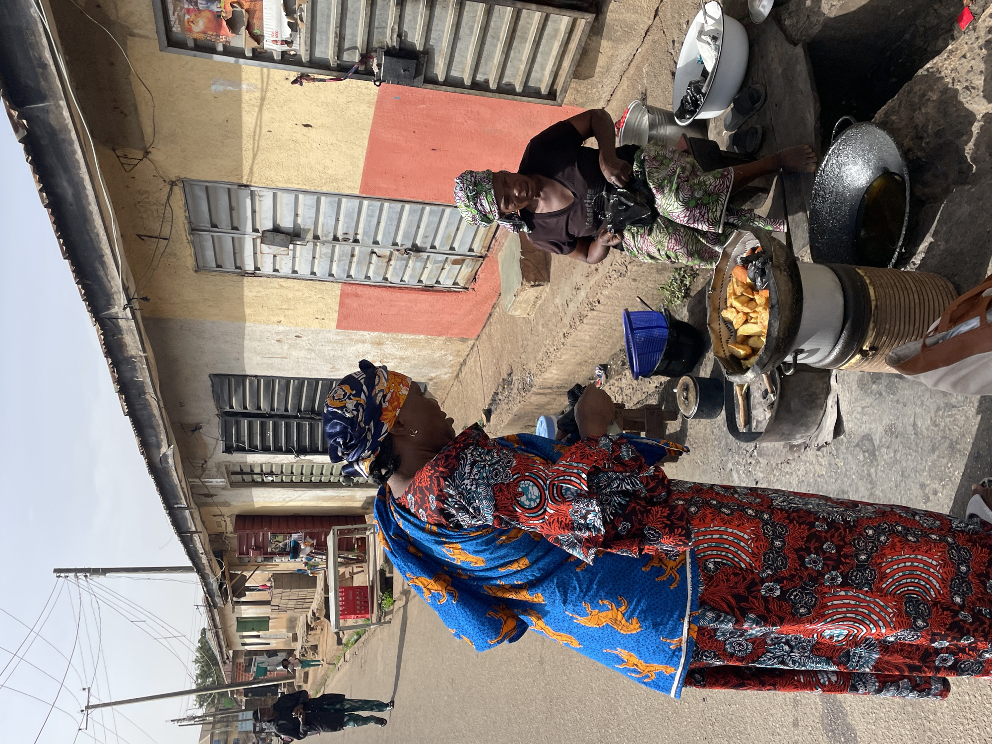 This image captures a bustling outdoor street scene, likely a food vending operation, in Jos, Nigeria, under bright, clear daylight. The sun casts strong shadows, suggesting midday or early afternoon.
Two women are prominently featured. In the foreground, a woman stands facing right, adorned in a vibrant, multi-patterned blue and red dress or wrapper, intricately decorated with yellow animal motifs, possibly leopards. She also wears a matching headwrap and a small earring.
Behind her, to the right, a second woman is seated on the ground, actively engaged in preparing food. She wears a green, white, and purple patterned wrapper, a dark t-shirt, and a colorful headwrap, and is barefoot. She is positioned next to an improvised cooking station that includes a large metal pot filled with golden-brown fried food, resembling potato chips or French fries, resting on a substantial cylindrical stove structure. Adjacent to this is a dark, round wok or pan, likely containing cooking oil. Several other kitchen items, such as a white enamel basin, metal buckets, and a blue plastic basket, are scattered around her on the dirt and concrete ground.
In the background, a building with sections painted in yellow, orange-red, and grey features multiple windows with corrugated metal shutters. Utility poles and power lines extend into the distance along a street visible on the left side of the frame. The overall scene depicts an active street market environment focused on local food preparation and sales. No discernible text is visible on any object or surface.