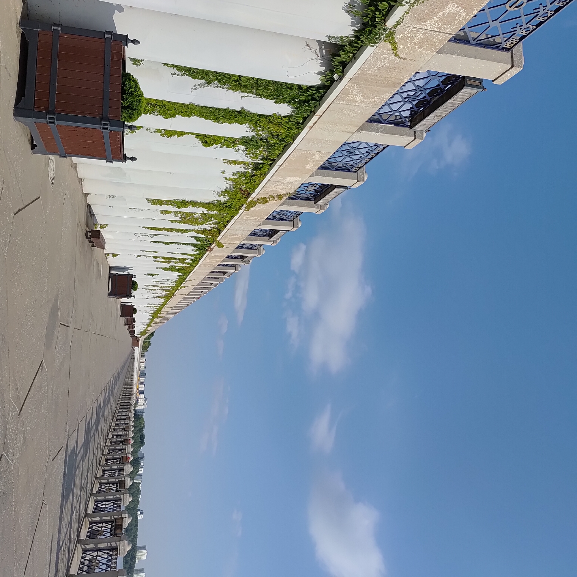 The image depicts a wide concrete embankment along a body of water, likely the Belaya River, in Ufa, Russia. The embankment is lined with a decorative metal fence and a stone wall. Green vines cascade down the stone wall in a regular pattern. Several large wooden planters with small green plants are positioned along the embankment, as well as a few small wooden benches. The pavement of the embankment is composed of large concrete slabs. In the distance, across the water, is a cityscape with numerous buildings and a line of trees. The sky is clear blue with a few scattered white clouds. The scene appears to be tranquil and well-maintained, suggesting a public park or recreational area. There are no people visible in the image. The sunlight casts distinct shadows from the fence onto the embankment, indicating a bright, sunny day.