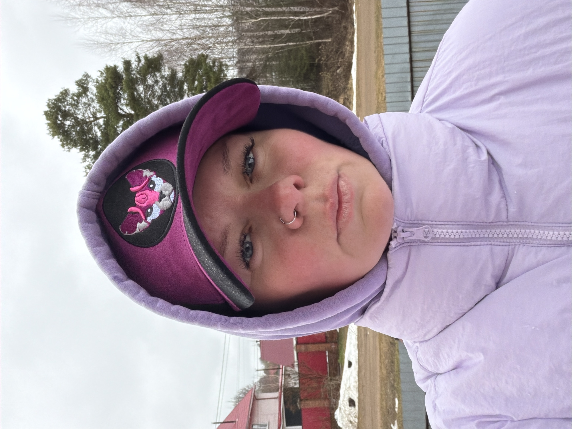 A person wearing a light purple hooded puffer jacket and a pink baseball cap with a black patch depicting a cartoonish animal is centered in the frame. The person has fair skin, blue eyes, and a silver nose ring. The hood of the jacket is up, framing the face and partially covering the cap.
The background shows a rural or suburban landscape. In the mid-ground, there is a dirt road, and beyond that, red and white buildings are visible. In the far background, there are bare trees suggesting either winter or early spring. The sky is overcast and grey.
It is not possible to determine the location as the city of Chaikovsky, Russia based solely on this image.
