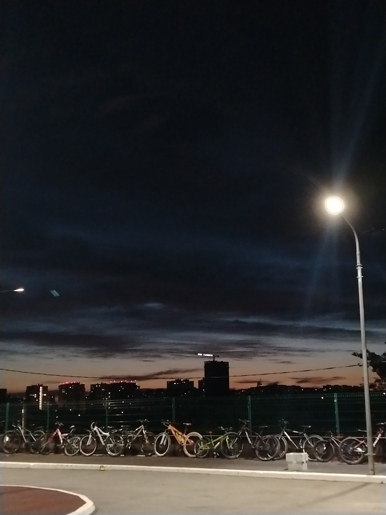 The image captures a row of parked bicycles under streetlights, with a city skyline faintly visible against the twilight sky. The bicycles are varied in style and color, suggesting individual ownership. A green fence separates the bicycles from the urban background. The sky displays dark clouds tinged with orange and pink hues from the setting sun. Based on identifying characteristics, the location appears to be Izhevsk, Russia. The scene suggests a quiet, possibly public, space at dusk.