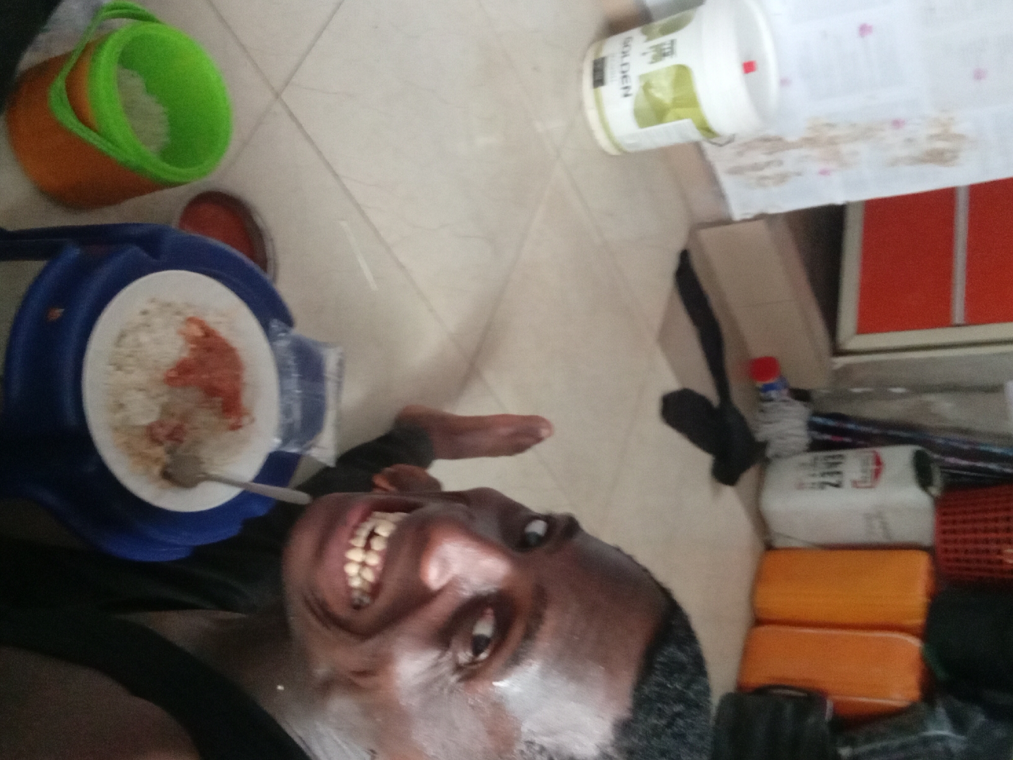 A medium shot depicts a dark-skinned man, close to the floor, tilting his head back towards the camera, revealing his teeth in a wide smile. He is wearing a black shirt or garment. To his left, a white plate containing rice and a reddish sauce sits on a blue plastic bowl or tray. A spoon rests on the plate. Above the plate, in the upper left corner, there are orange and green plastic buckets. To the right of the man, in the upper right corner, is a white can of paint labeled "GOLDEN" and a cardboard box sits on top of a white cloth. Further to the right, there is a collection of various items, including a bottle, orange sponges, a basket, and other indiscernible objects. The floor is tiled with light-colored, square tiles. The location is specified to be Akwete, Nigeria.