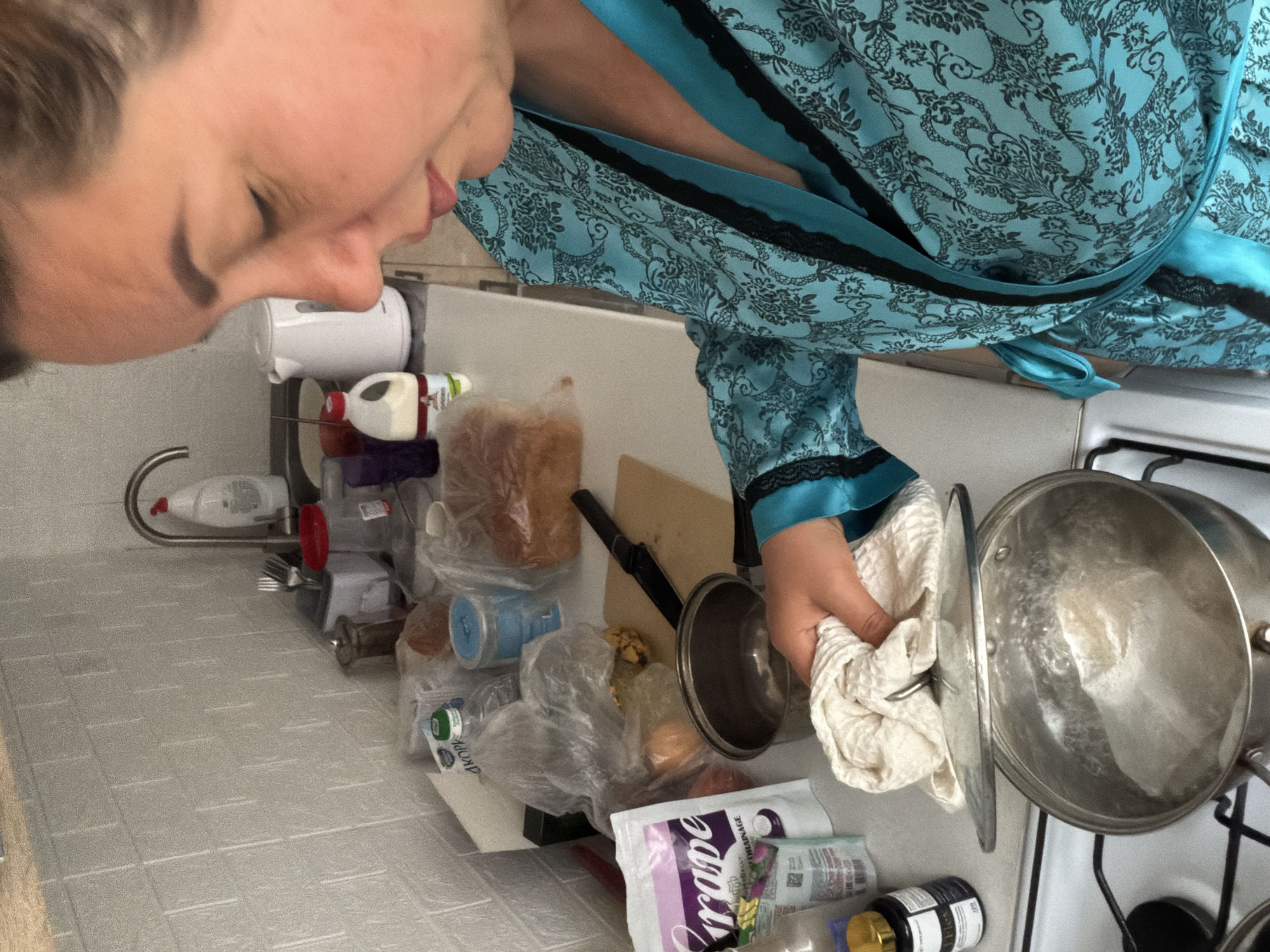 A woman wearing a blue and black patterned robe stands in a kitchen. She is holding a pot lid with a white towel. Two pots are on a white stove top. The countertop is filled with various items, including packaged food, jars, and kitchen utensils. The location is Leningradskaya, Russia, as indicated by Cyrillic text on one of the packages on the counter.