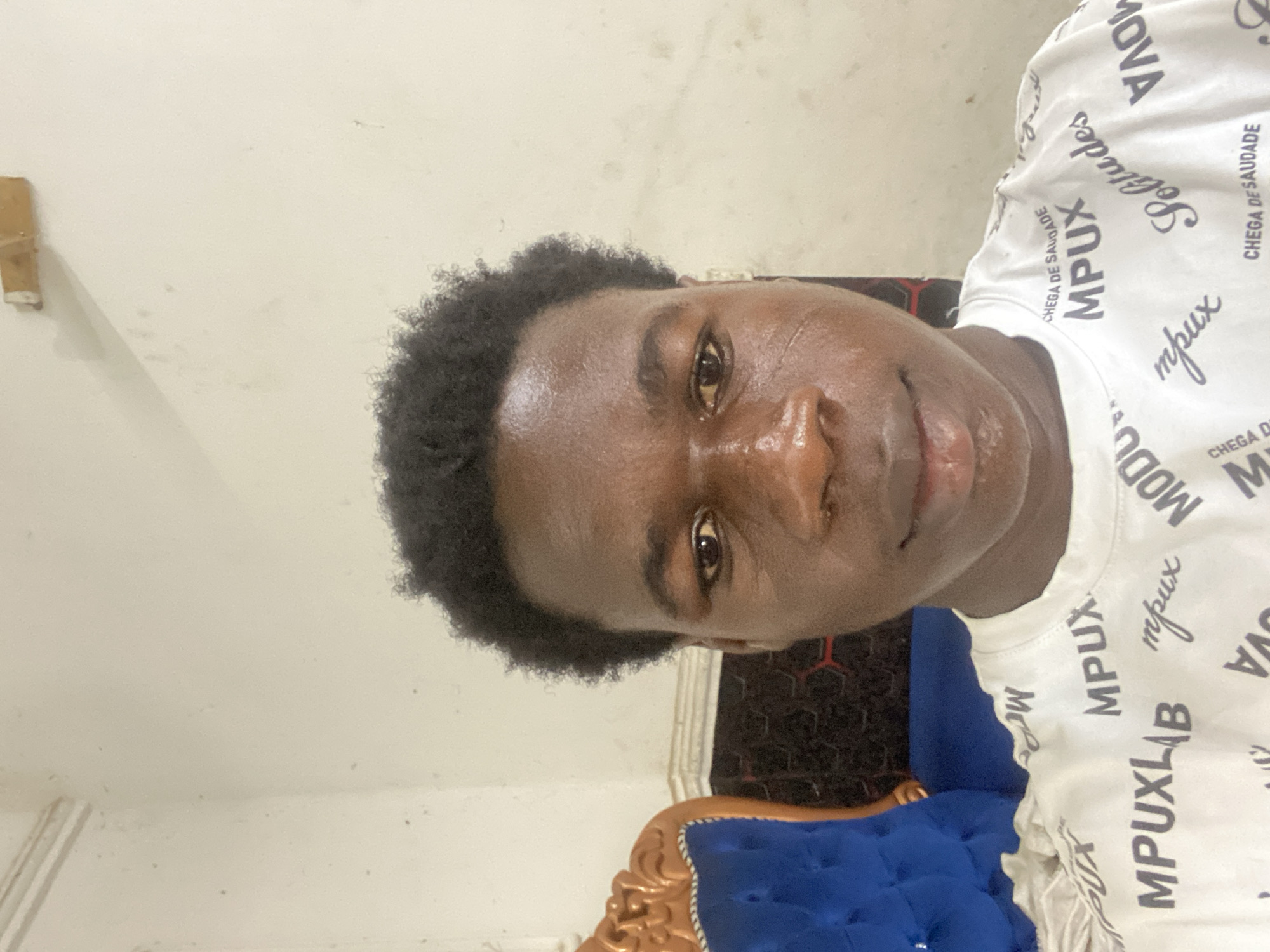 The image shows a young man with dark skin and short, curly black hair. He is wearing a white t-shirt with the word "MPUX" printed repeatedly in a dark font. He is looking at the camera with a slight smile. Behind him is a blue, tufted couch with gold trim. The background is a plain, light-colored wall. The location is unknown as the image contains no information to identify it as Mararaba, Nigeria.