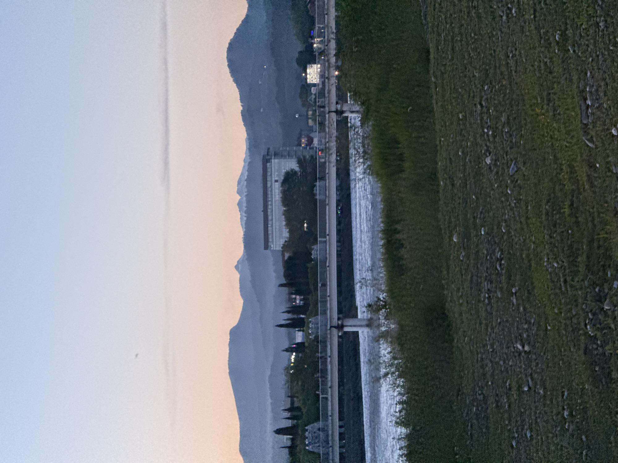 The image captures a serene landscape view, likely at dusk or dawn, across a body of water in Sochi, Russia. The scene is horizontally composed, with a grassy riverbank occupying the right side of the foreground. This bank is dotted with small stones and has varying textures of grass.
To the left of the grassy bank, a wide river or canal flows, its surface reflecting the ambient light and showing ripples. Spanning across this water body in the midground is a long, light-colored structure, possibly a pedestrian bridge or pier, supported by pillars submerged in the water. This structure features railings running along its length.
Beyond the water and the structure, the far bank is lined with a mix of evergreen and deciduous trees, appearing as dark silhouettes against the lighter sky. Several buildings are visible among the trees and further along the bank. Some buildings show illuminated windows, indicating artificial light sources are active, consistent with twilight hours. One particularly large, modern building with a distinct architectural style stands out among the structures.
In the background, successive layers of mountains rise, appearing hazy and bluish-grey, suggesting distance and the soft light conditions. The sky above these mountains transitions from a pale blue at the top to a soft, warm orange-pink closer to the horizon, reinforcing the impression of either sunrise or sunset. A faint, thin vertical streak, possibly a contrail from an aircraft, is visible in the upper sky.
No people or specific activities are visible in the image, contributing to a calm and tranquil atmosphere. The overall impression is one of natural beauty blended with urban development, characteristic of a mountainous coastal city like Sochi. There is no visible text.