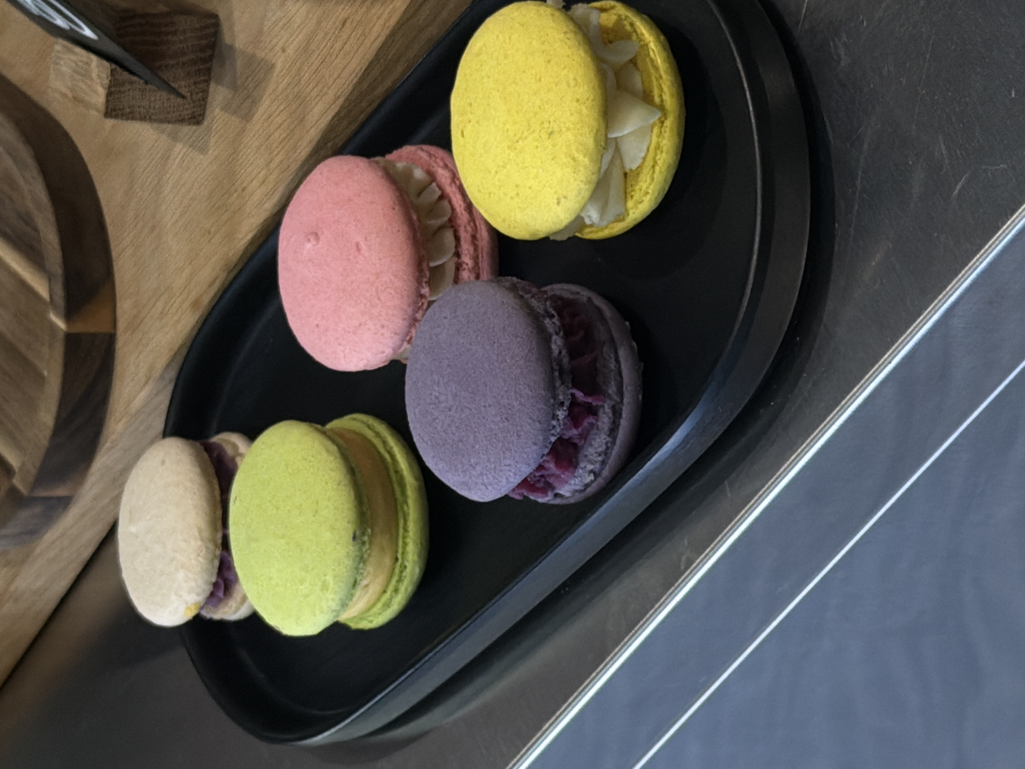 A black tray on a table holds five macarons of different colors: yellow, pink, purple, green, and white. The yellow macaron appears partially open. The tray sits on a wooden table and a metal surface. There are no people visible in the image. There is no explicit information that confirms this image was taken in Belgrade, Serbia.