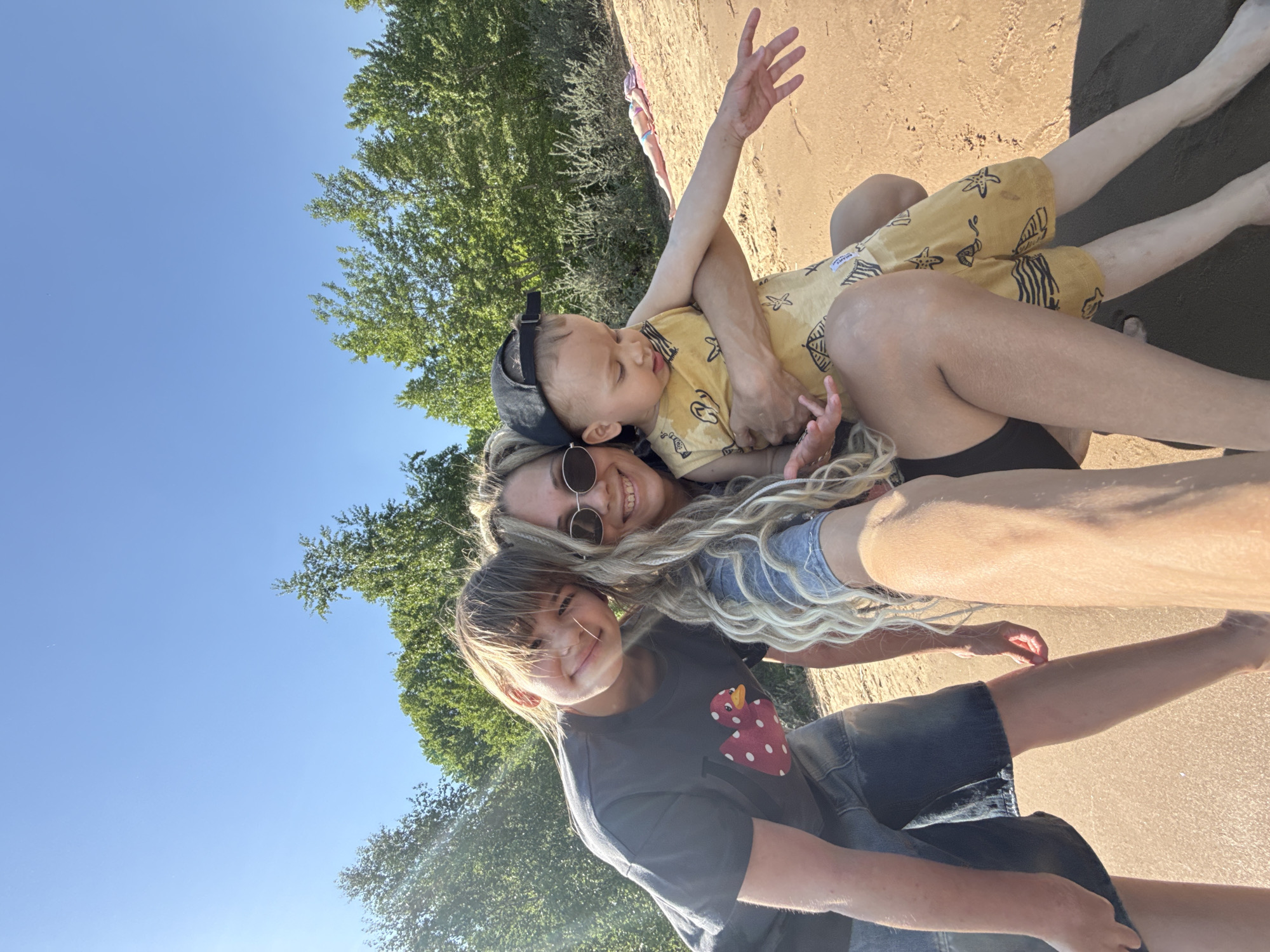 The image depicts a woman with blonde hair wearing sunglasses, a young child in a yellow patterned outfit and a cap, and an older child in a dark t-shirt, all lying on a sandy surface, likely a beach. Trees are visible in the background, along with a clear blue sky. The location is Unknown, Unknown.