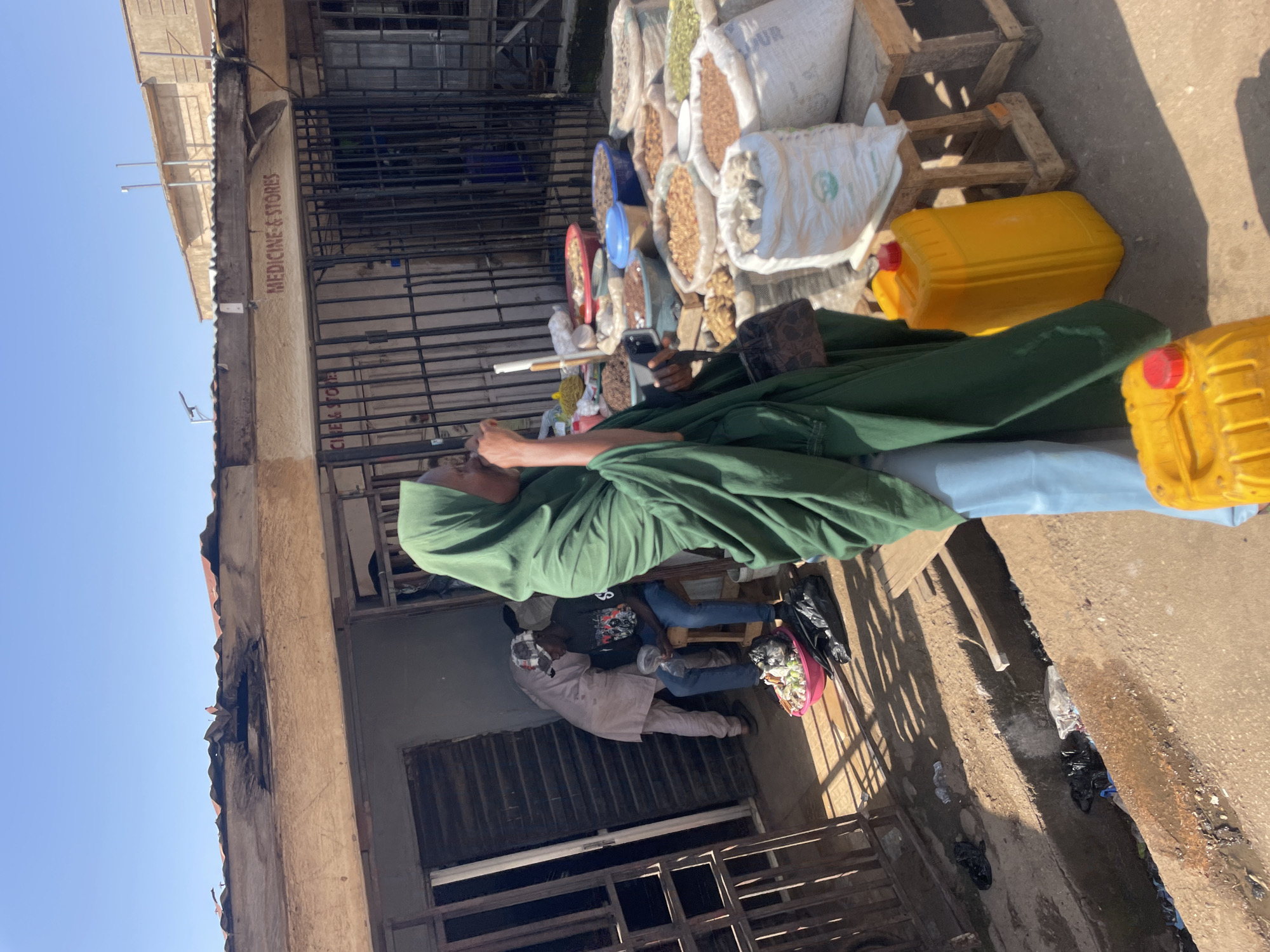 In an outdoor market scene, a woman wearing a green hijab and light blue trousers stands with her back to the camera, looking to her right. She holds a dark handbag and a mobile phone. In the foreground, two yellow jerrycans with red caps are visible. Behind the woman, a stall displays various sacks of grains and spices, some in woven bags and others in white plastic bags. A small blue bucket and several red bowls are also present. To the left, two men are interacting near a doorway. One man, wearing a checkered cap and dark t-shirt, sits on a stool in front of a pink bowl filled with various items and black plastic bags. Another man, dressed in a light-colored garment, stands beside him, partially obscured. The background shows a building with a corrugated metal roof and metal bars in front of its windows and doorways. A sign on the building reads "MEDICINE & STORES". The ground is paved with concrete, and a narrow, open drain filled with debris runs along the lower left of the image. The scene is illuminated by bright sunlight, casting shadows.