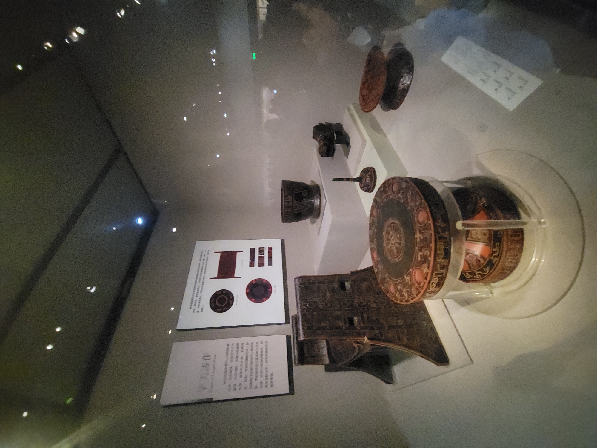 This image depicts an indoor exhibition display, likely at a museum, featuring ancient Chinese lacquerware. The scene is set within a dimly lit environment, with spotlights illuminating the artifacts and creating reflections on the glass display cases and the ceiling.
The display showcases several intricately decorated lacquerware items. Prominently featured in the foreground is a round, lidded box with elaborate patterns, supported by a clear acrylic stand. To the left, a tall, dark, intricately carved wooden object, possibly a seat or a stand, is partially visible. Behind these items, several other artifacts are arranged on white pedestals. These include a dark bowl with decorative motifs, a small sculpture resembling an animal head, a slender, knobbed object, and a shallow dish.
On the wall behind the artifacts, there are two display boards with Chinese text and illustrations. The board on the left, titled "Exquisite Lacquer Ware," provides descriptive information about the pieces. The board on the right features diagrams and images of various lacquerware objects, including circular plates and a rectangular piece that appears to be a bed or a platform.
Reflections of people and lighting fixtures are visible on the glass surfaces, indicating the presence of museum visitors and the exhibition's lighting design. The overall ambiance suggests a cultural or historical exhibition. While the specific museum or gallery is not identified, the presence of Chinese text and the nature of the artifacts strongly suggest a location in China. Given the request to assume a location in Qingyang District, China, this exhibition could be housed within a museum in that district.