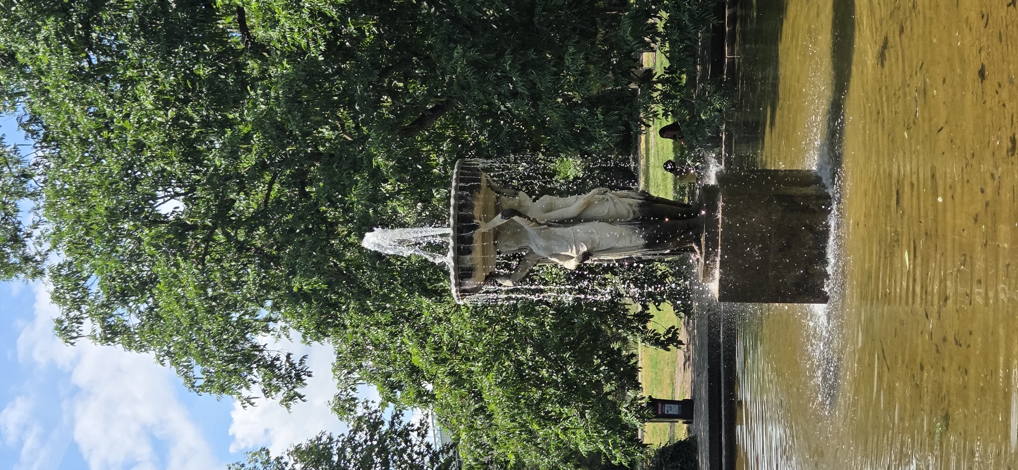 The image depicts a fountain in the Schlossgarten in Karlsruhe, Germany. The fountain features a circular basin supported by three female figures, with water spraying upwards and cascading down. The fountain is situated in a pond with murky, greenish-brown water. Surrounding the pond is a paved walkway and lush green grass, dotted with trees. In the background, two individuals are seated on the grass, partially obscured by foliage. To the left of the fountain, a black trash bin with a yellow lid is visible near a path. The sky above is blue with scattered white clouds, and the sunlight casts shadows on the scene.