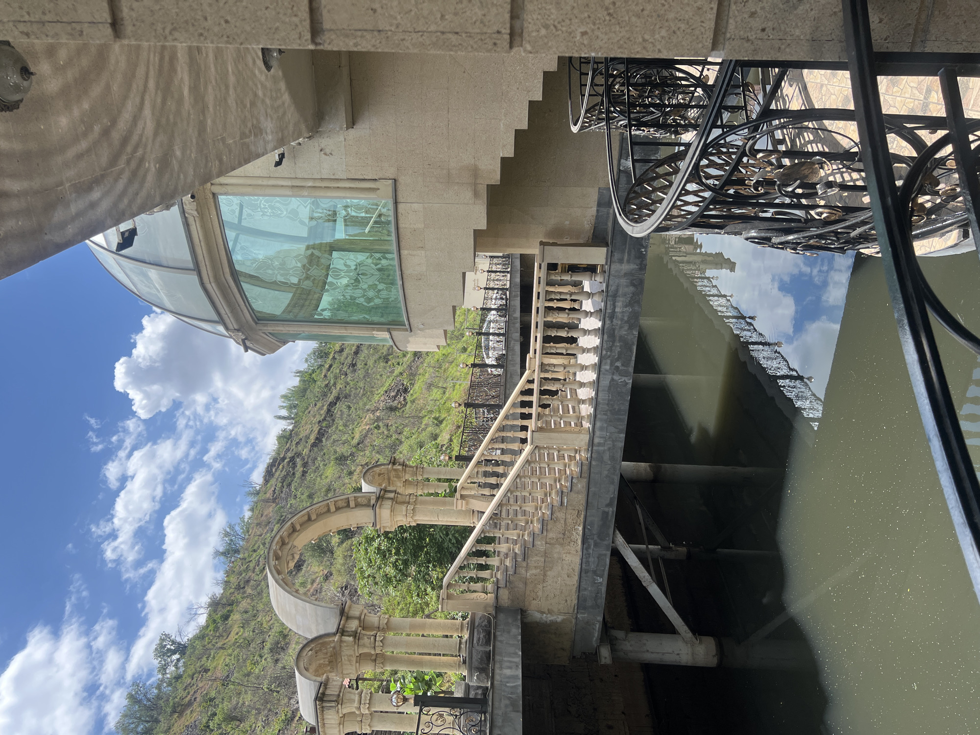 The image presents a multi-level architectural complex constructed from light-colored stone, situated next to a body of water and a steep, verdant hillside. The scene is bathed in bright daylight under a clear blue sky dotted with fluffy white clouds, characteristic of a sunny day in Yerevan, Armenia.
The building incorporates classical design elements, including elegant stone staircases with balustrades that descend towards the water. Arched passageways and columns add to the grand aesthetic. A prominent feature is a curved, possibly domed, glass section on one part of the building, offering wide views. A small black security camera is visible on its exterior. Further details include intricate black wrought iron railings with decorative scrollwork, some incorporating stylized bird motifs, which line walkways and overlook the water.
The body of water reflects the sky and parts of the surrounding structures. Its color appears somewhat murky or greenish-brown in close-ups, with light glinting off its surface. Support beams and pipes are visible beneath the structures, extending into or across the water. The background is dominated by a lush green hillside with scattered trees.
No people are present, leaving the scene peaceful and unpopulated. On one of the building's walls, a small rectangular sign is partially visible, with text that appears to read "Ristorante" or similar, suggesting the complex may be a dining establishment, hotel, or a recreational venue. Light fixtures are also visible on the stone walls.