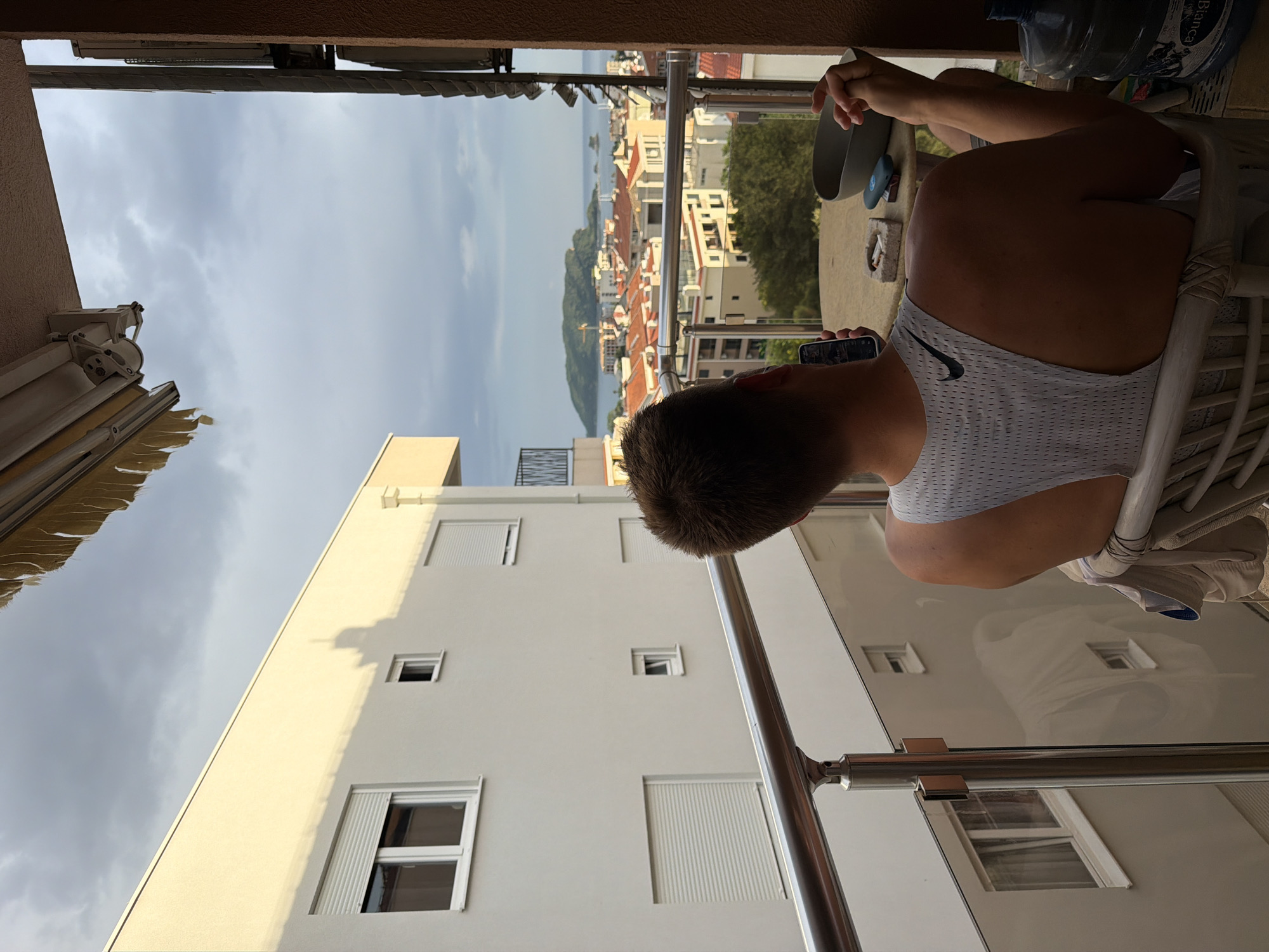 A man wearing a white Nike athletic shirt sits in a white wicker chair on a balcony, holding a phone to his ear. In the background is a scenic view of Budva, Montenegro, featuring buildings with red tile roofs, green trees, and a body of water under a cloudy sky. The balcony has a stainless steel railing. A white building with multiple windows is visible to the left. Bottles of water are on the table next to the man.