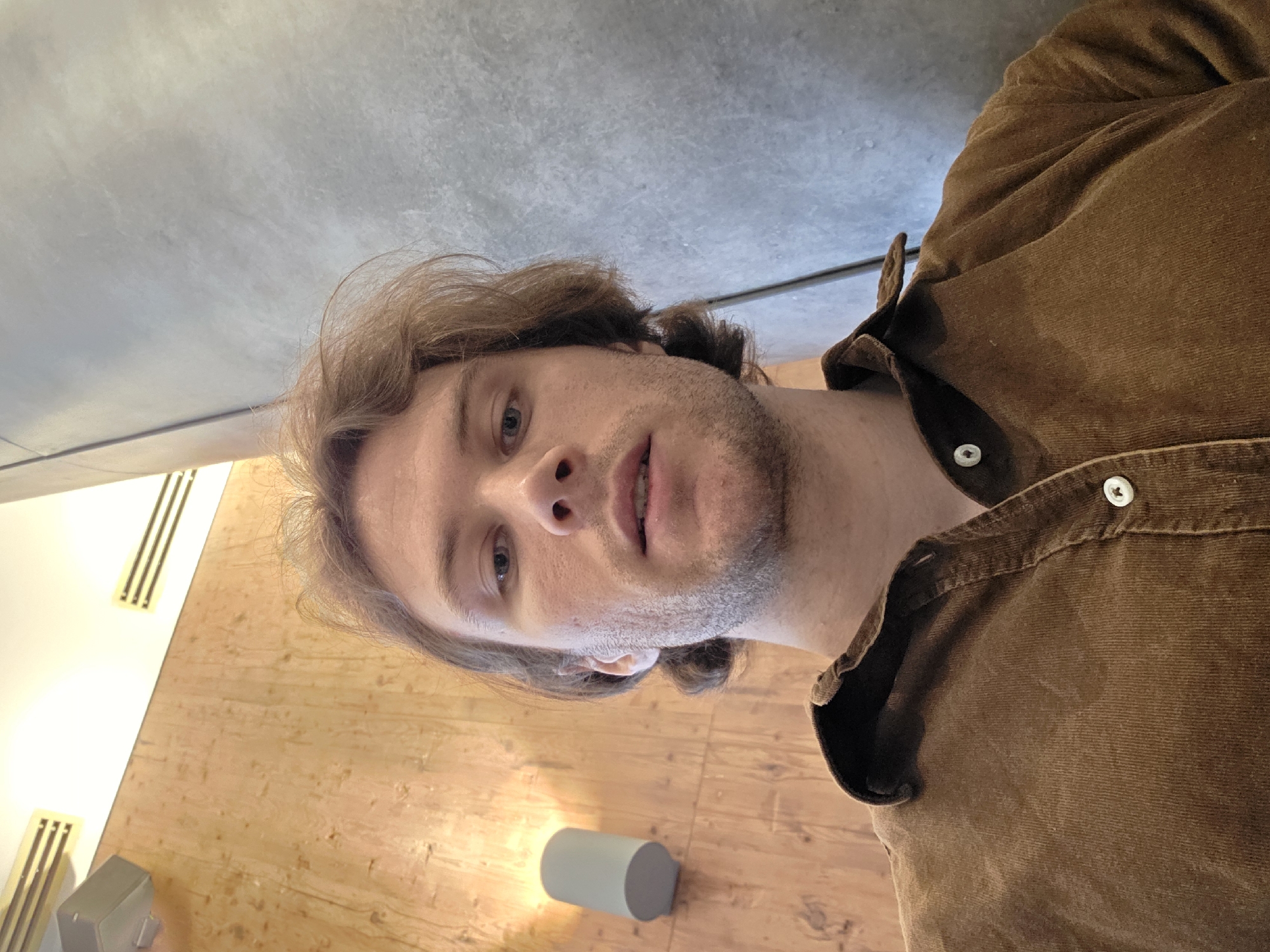 A selfie-style photograph depicts a fair-skinned male with light brown hair and stubble, wearing a brown corduroy shirt. The background consists of a light brown wood-paneled wall with vertical ventilation slats and a light gray concrete ceiling. A small gray cylindrical object is partially visible on the wood surface. No specific landmarks or elements clearly indicate a location in Batumi, Georgia. Therefore, based solely on the visual information, the city cannot be confirmed.