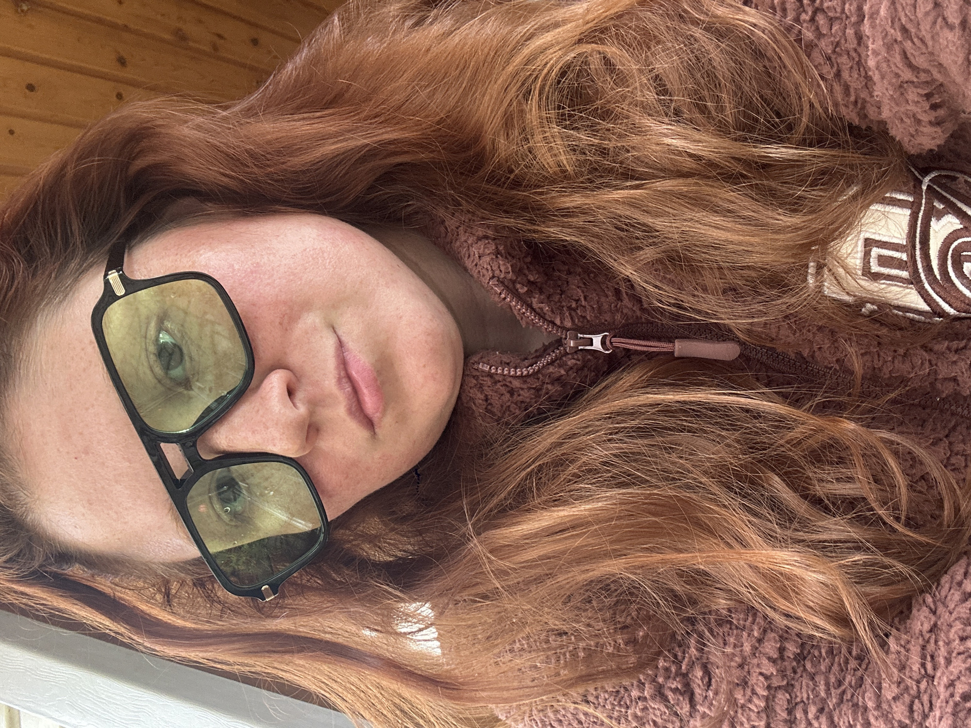A woman with long, auburn hair lies on her side wearing black, rectangular glasses with green lenses and a brown, fleece zip-up jacket. The top of a light-colored wall is visible in the lower-left corner. There is no evidence of Nizhny Novgorod or any other specific location.