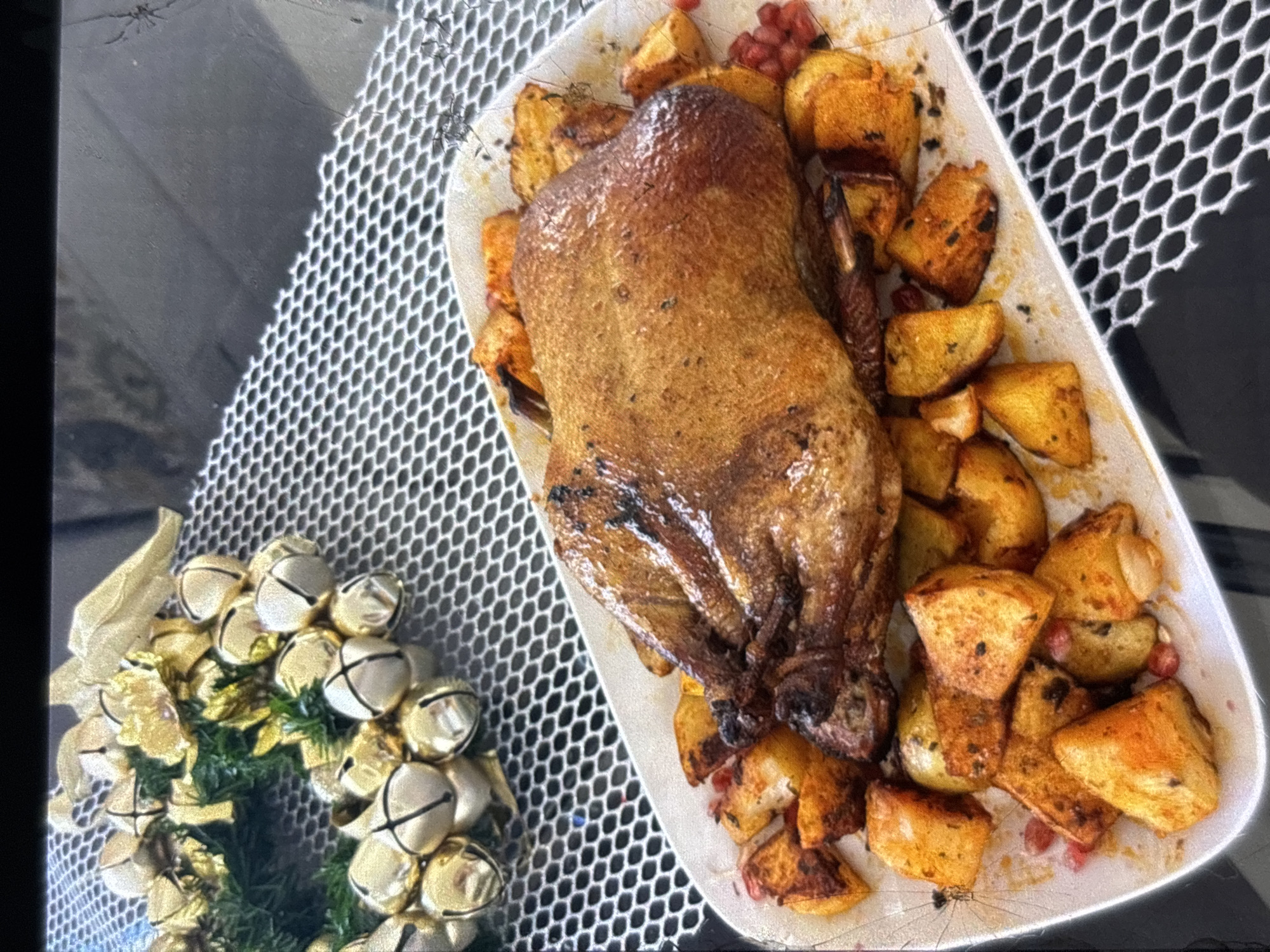 A whole roasted duck, golden brown and glistening, rests on a white platter surrounded by roasted potatoes and scattered pomegranate seeds. In the upper left corner, a festive wreath adorned with gold bells and greenery is visible. The platter is placed on a black surface with a honeycomb-like texture. The scene appears to be set indoors, likely for a meal or celebration. The image does not provide any information about the city of Orihuela, Spain.