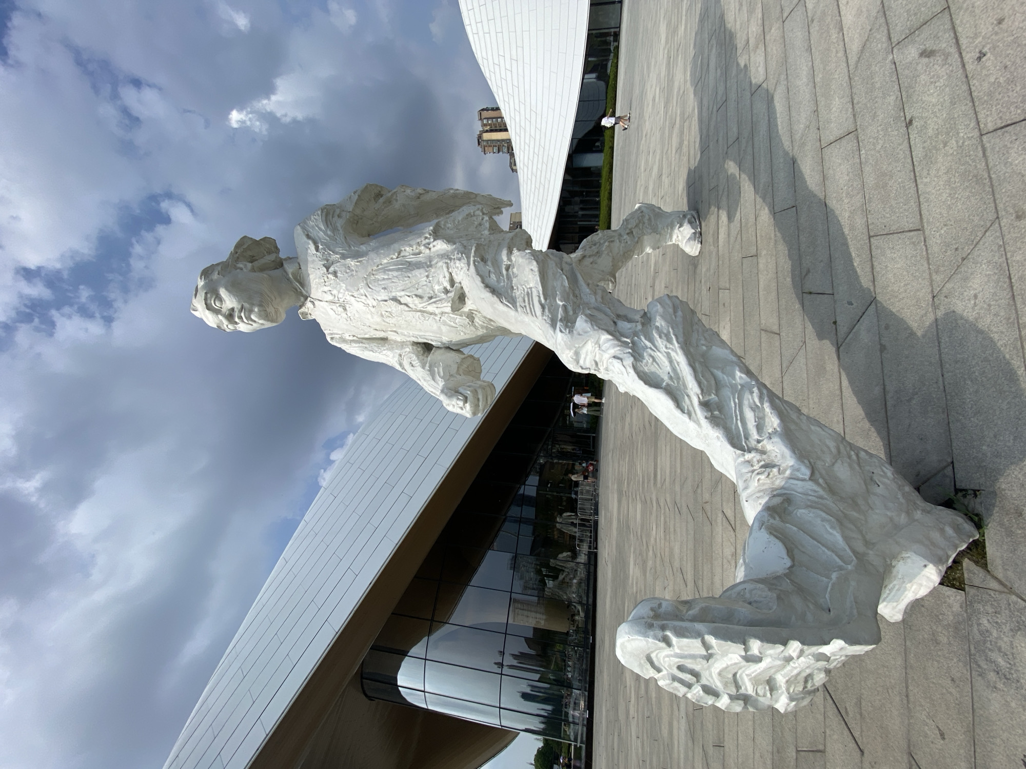 A white, abstract sculpture of a person in mid-stride is positioned in the foreground of an outdoor plaza. The sculpture is textured and appears to be made of a rough material. The figure's left leg is extended forward and downward, touching the ground, while the right leg is bent and raised behind. Behind the sculpture, a modern building with a white, undulating roof and a large glass facade is visible. The sky is partly cloudy, with dramatic dark clouds mixed with patches of blue sky. The plaza is paved with gray stone tiles. In the background, a few people are walking in the distance, and residential buildings can be seen beyond the modern structure. The scene suggests a public space, possibly near a cultural institution or landmark in Jinniu District, China.