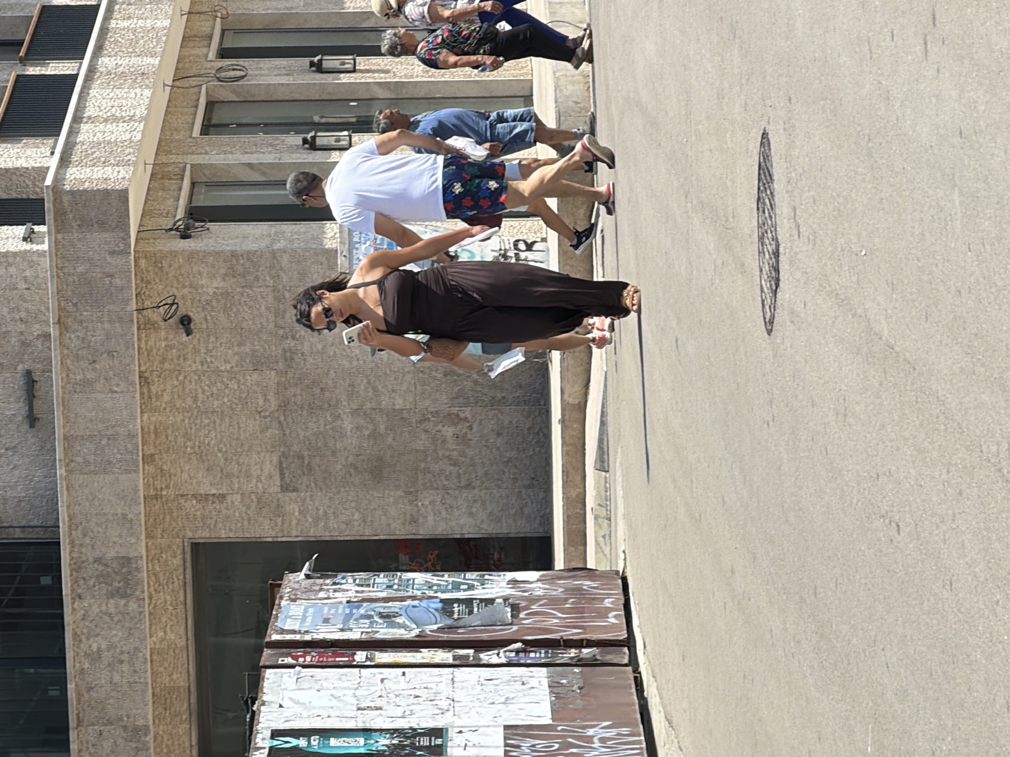 The image depicts a street scene in Budva, Montenegro, under bright daylight. In the foreground, a grey asphalt road is visible, with a circular manhole cover in the lower center. To the left, a weathered, graffiti-covered metal structure, possibly a kiosk or information board, stands at the edge of the sidewalk.
Several people are walking on the sidewalk. Closest to the camera, a woman with long dark hair and sunglasses is wearing a dark brown sleeveless jumpsuit and sandals. She is holding a smartphone in her left hand and a small brown purse on her left shoulder. Behind her and to the right, a man in a white t-shirt and floral-patterned shorts walks away from the camera. Further back, other individuals are visible, including a man in a blue shirt and denim shorts, and an older woman with grey hair wearing a colorful patterned top and dark pants. The background features a building with a textured beige facade and dark-framed windows, some with black railings. The overall scene suggests a bustling urban environment on a sunny day.
