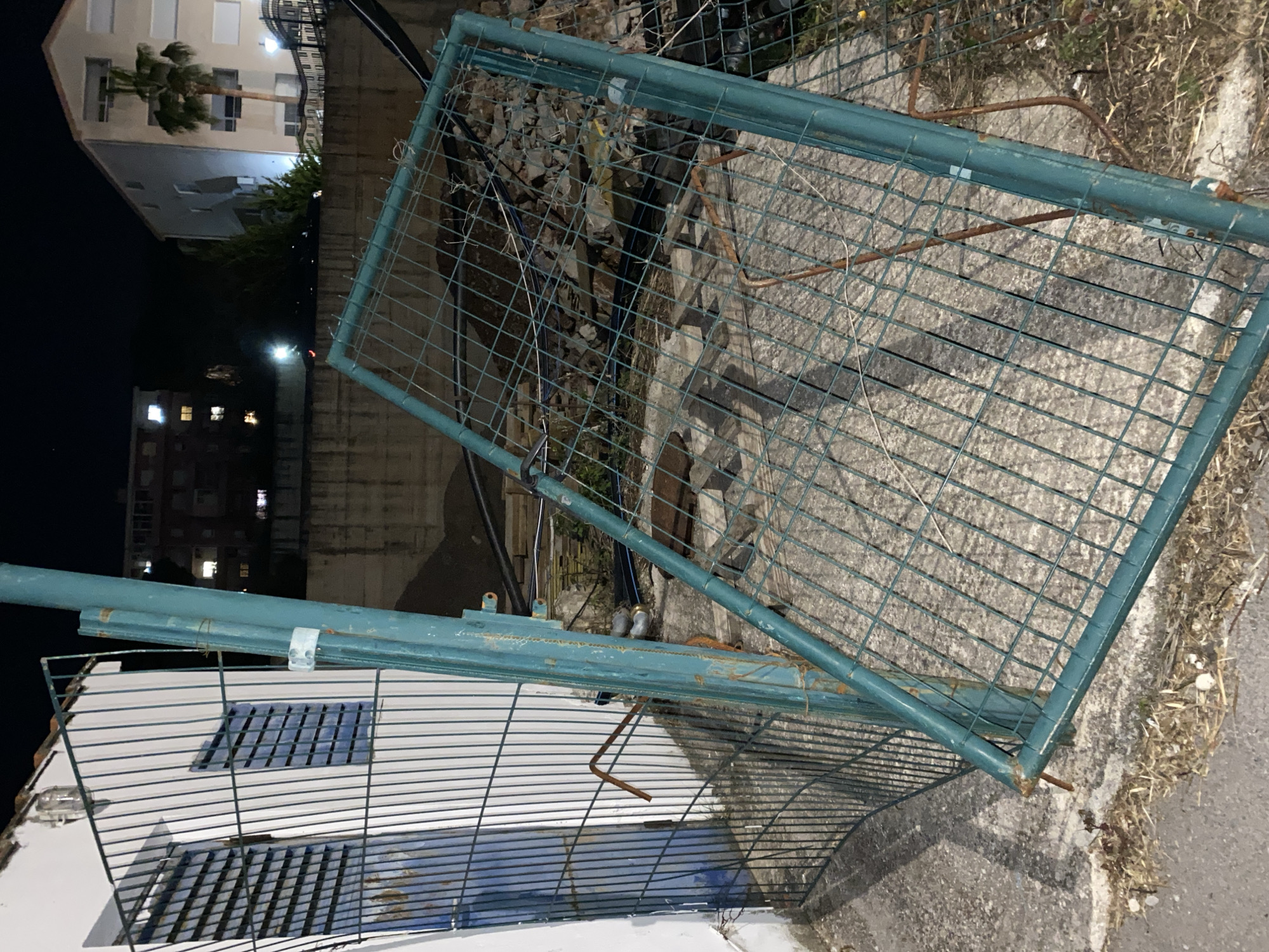 The image depicts a haphazard scene likely in the city of Budva, Montenegro, given the building style and vegetation visible in the background. Discarded or damaged metal fencing is the main subject, including a larger, rectangular green-painted section with a wire mesh infill, and a smaller, horizontal section with a similar design lying on top of a white structure with blue-shaded windows. Debris, possibly from construction or demolition, is visible behind the larger fencing section. The environment suggests an urban area, with multi-story buildings illuminated by streetlights in the distance against a night sky. The scene appears to be a roadside or vacant area, possibly near a construction site.