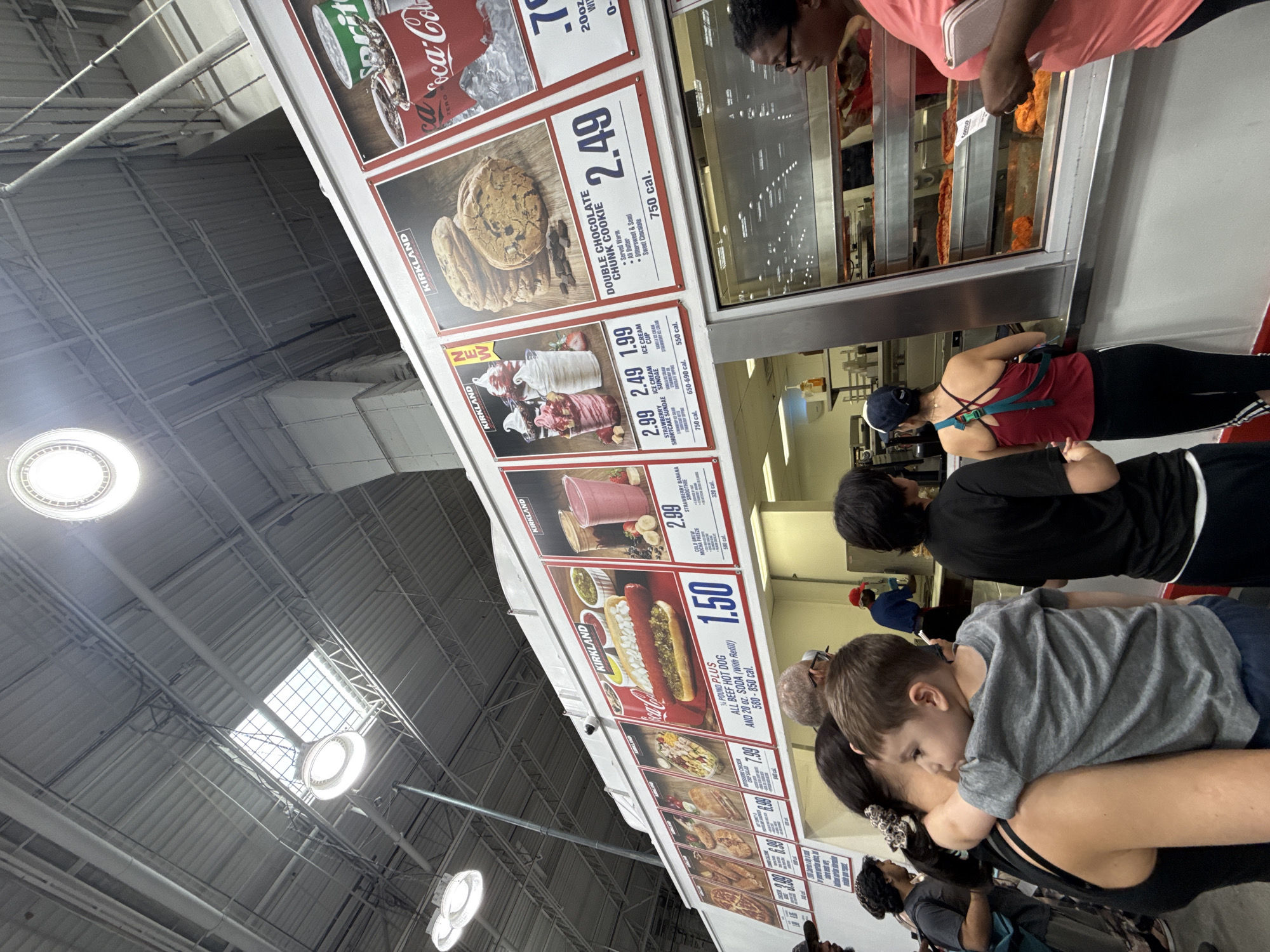 This is a medium-shot, wide-angle photograph of a Costco food court with a visible menu board and a line of customers. The environment appears to be indoors, with a high ceiling and industrial lighting. The menu board displays various food items, including hot dogs, ice cream sundaes, cookies, and smoothies, with prices and calorie information. The customers are a diverse group of people of various ages and ethnicities, some are looking at the menu, some are in line, and some are interacting with each other. A young child is being held by an adult in the foreground. Behind the counter, food service workers are visible, preparing food. The overall scene depicts a typical day at a Costco food court. There is no specific information or visual cues in the image to indicate the location is in the City of Yonkers, United States of America.