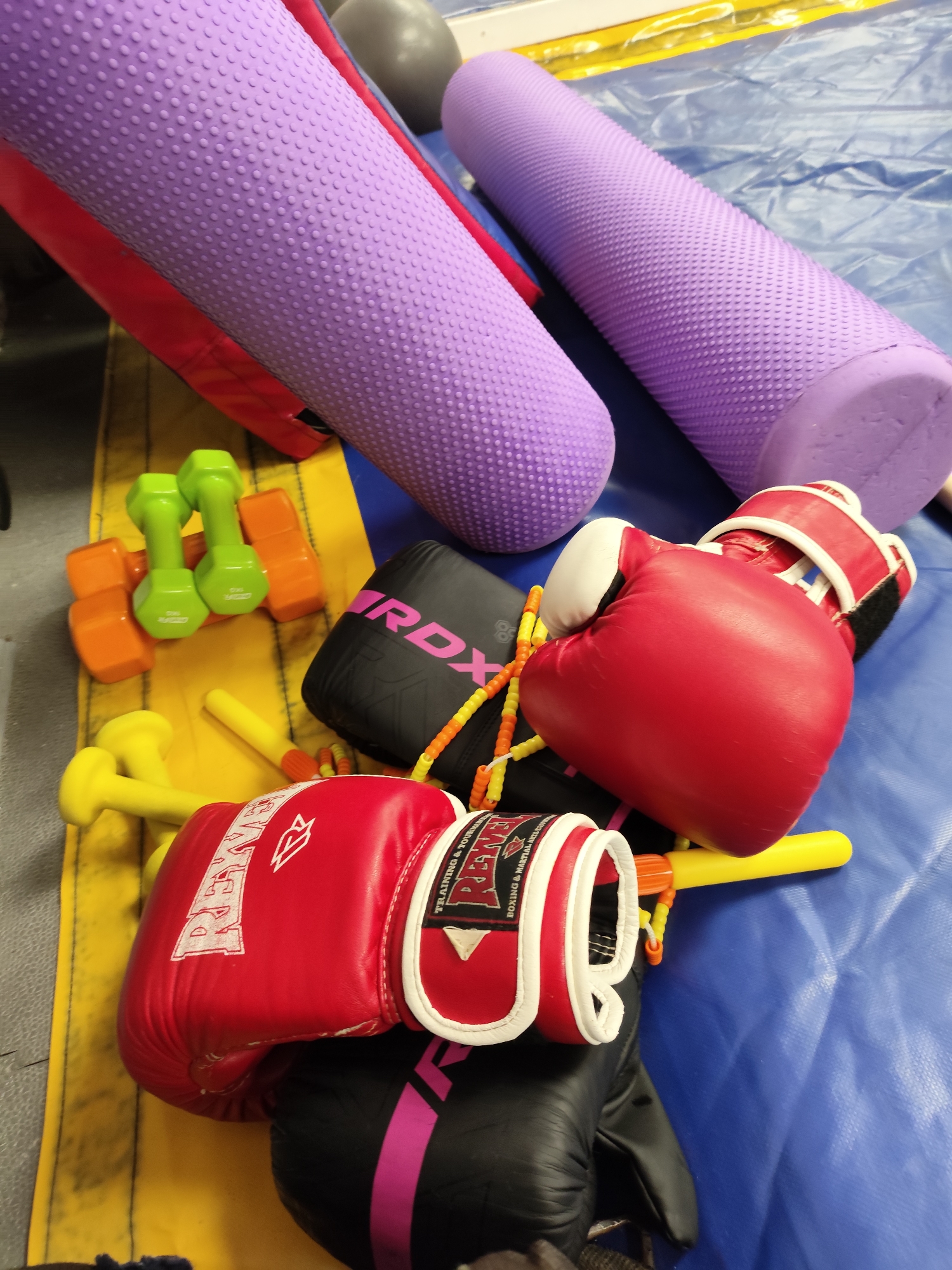 In Krasnodar, Russia, a close-up shot captures a collection of boxing and fitness equipment arranged on a blue mat. Two purple foam rollers, one partially obscured by a red object, are positioned diagonally across the frame. To the left, two pairs of dumbbells, one green and one orange, rest on a yellow mat. Below them, yellow handgrips are visible, along with a black RDX bag. A pair of red boxing gloves is prominently displayed, with one glove on top of a black bag. A jump rope with orange and yellow beads is also present. The scene is static, with no people or active interactions. The lighting is even, and the focus is sharp, highlighting the textures and details of the equipment.