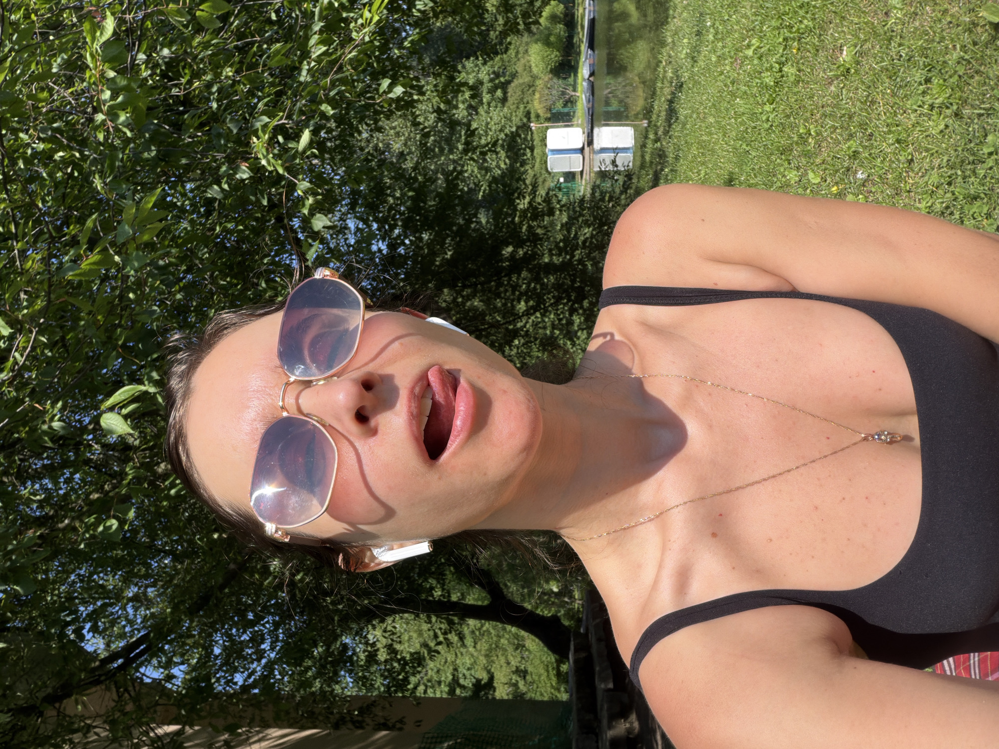 A woman with her tongue sticking out is captured in a selfie outdoors. She is wearing sunglasses, white earbuds, and a black tank top with a necklace. The background features lush green trees, a body of water, and two portable toilets on a distant shore. The scene appears to be a park or recreational area with a lake, likely in Saint Petersburg, Russia, given the typical summer vegetation and landscape.
