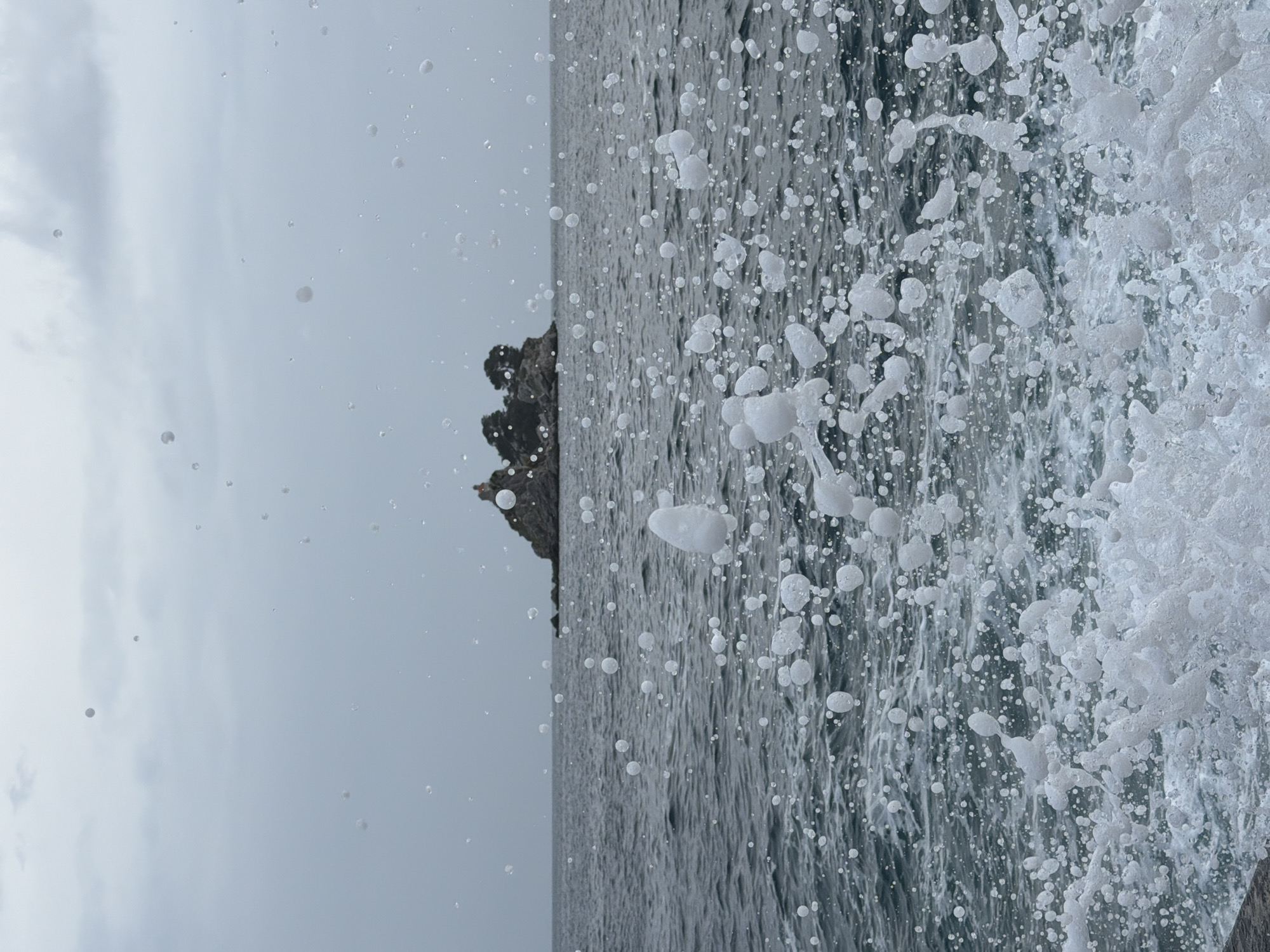 A sideways, close-up shot of water crashing against the Petrovac centar coastline in Montenegro. The right side of the image is filled with white sea foam from the waves. The water is grey and choppy. A rocky outcrop is partially visible on the left side of the frame. The sky is overcast and pale. Small droplets of water are suspended in the air.