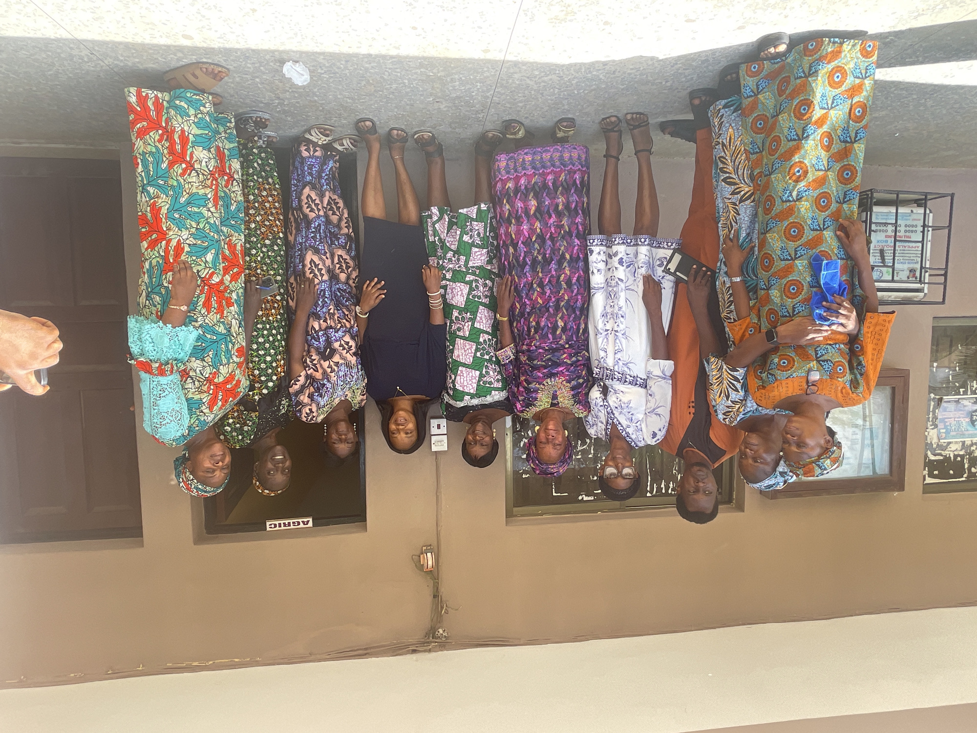 A group of nine people stands shoulder to shoulder in front of a building in Badagry, Nigeria. All are facing the camera and appear to be posing for a photograph. Eight are women, and one is a man. The women are wearing colorful, traditional African dresses. The man is wearing an orange shirt. A sign labeled "AGRIC" is visible in a window behind them. The building appears to be made of concrete and is a light tan color. Windows are visible behind the group, and a electrical outlets are visible on the wall.