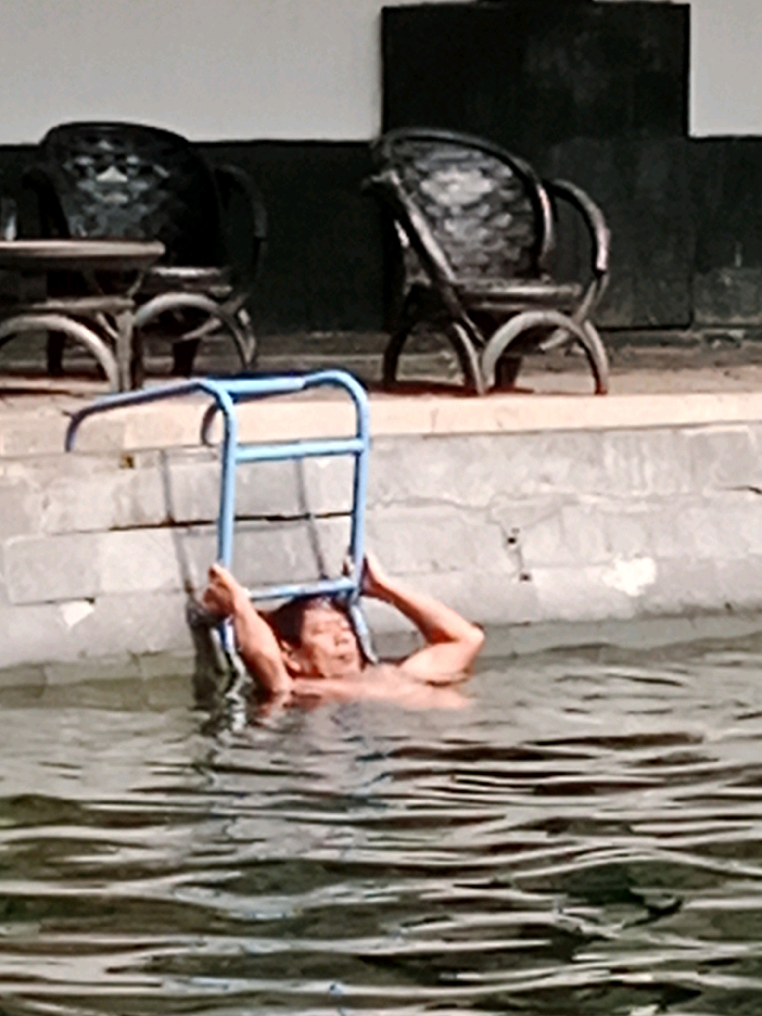 The image shows a person partially submerged in a body of water, holding onto a blue ladder. The person's head and shoulders are visible above the water, and they appear to be in a relaxed or resting position. The ladder is a pool ladder, suggesting the person is in a swimming pool. In the background, there are two chairs, likely outdoor furniture, positioned near a concrete wall or edge. The environment appears to be an outdoor setting, possibly a patio or poolside area. The lighting suggests it is daytime. There is no indication of the specific location within Jentekan, Indonesia.