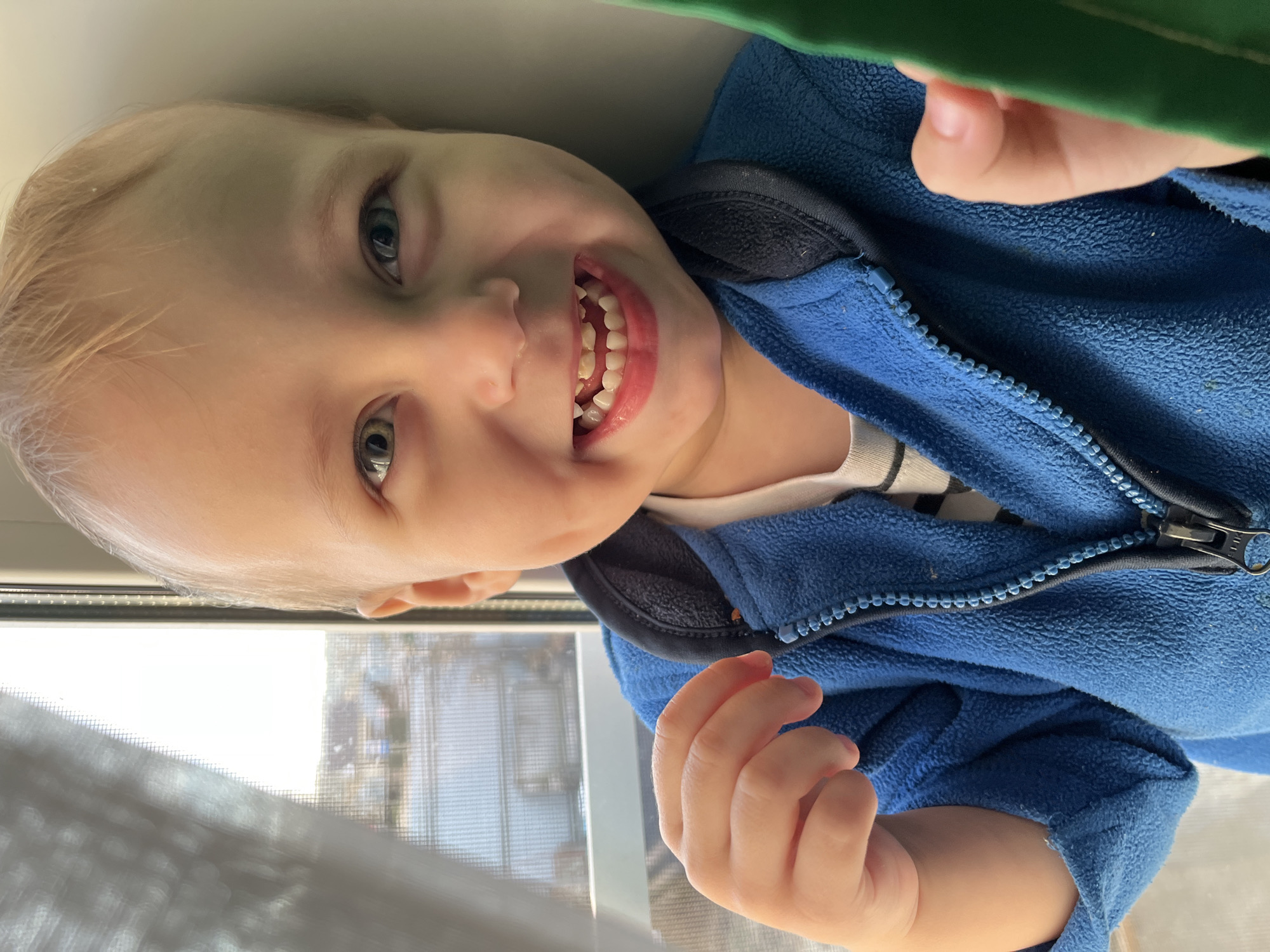 A young child with blonde hair and visible teeth is smiling. The child is wearing a blue zippered fleece jacket over a white shirt with black stripes. The child's hands are visible. The background includes a window with a screen and a glimpse of an outdoor scene with buildings. The location is unspecified, and there is no indication of the city of Фоки, Russia.