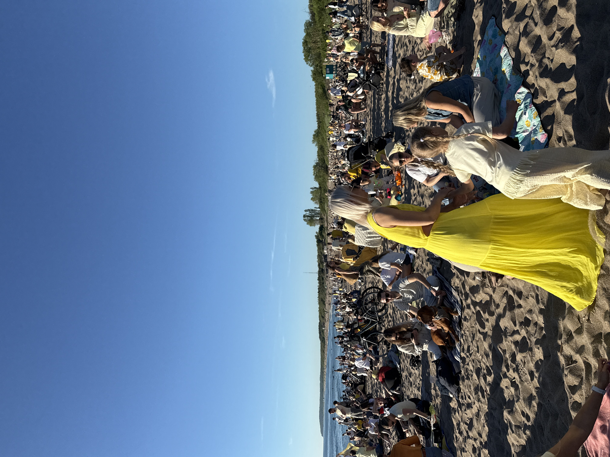 A wide-angle, eye-level outdoor shot shows a crowded beach scene on a sunny day with a clear blue sky. A large group of people are scattered across the sandy beach, many sitting on blankets or towels. Some are gathered in small groups, appearing to socialize or relax. A few individuals are standing, and children are visible among the crowd. Several strollers and bicycles are present, suggesting a leisurely outing. The beach extends into the distance, meeting the ocean, with a faint line of trees visible along the coastline. The overall atmosphere is one of a busy, communal gathering on a warm day.