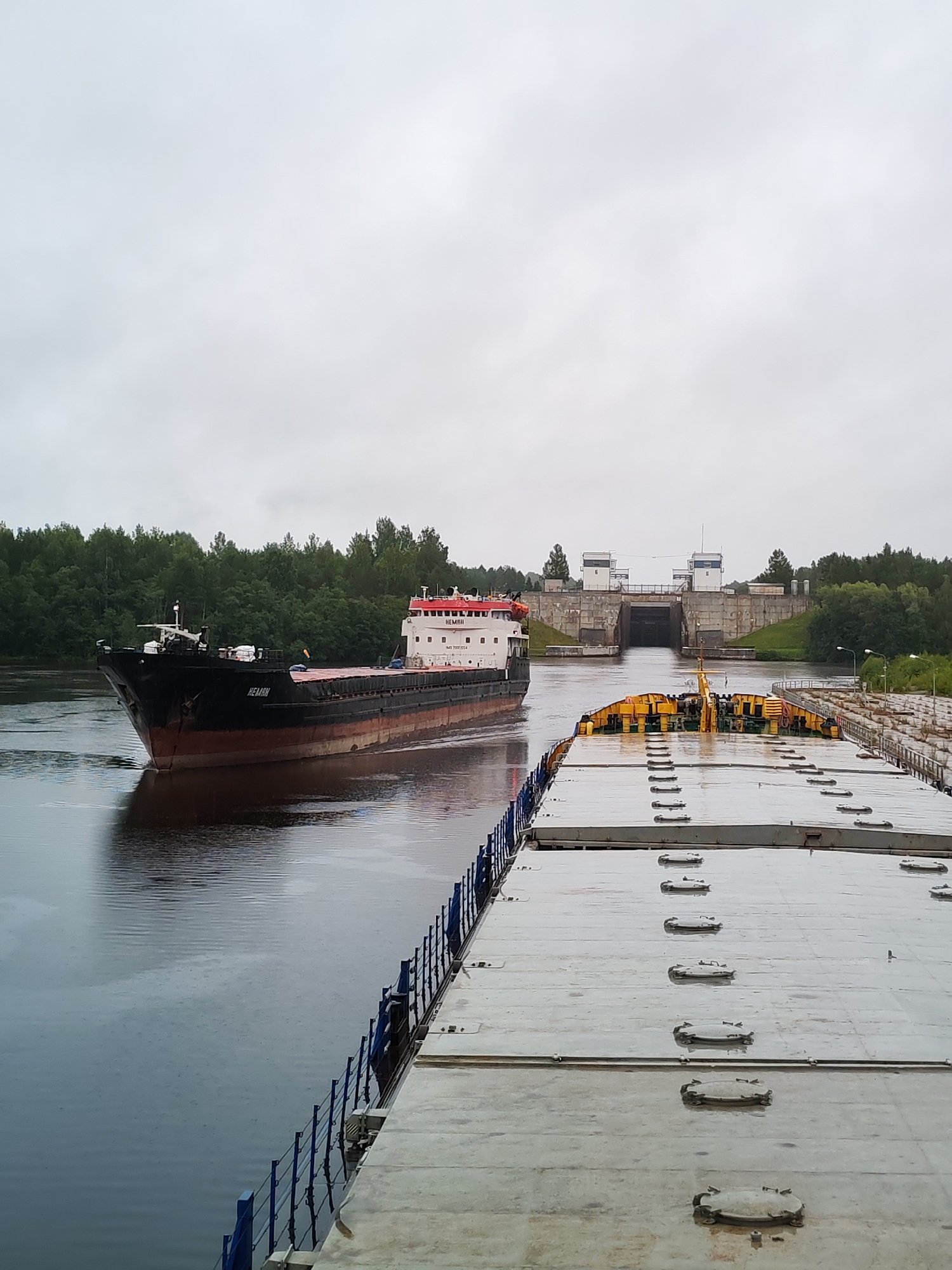 The image captures a scene on a canal in Devyatiny, Russia. In the foreground, a large, flat-topped barge dominates the right side of the frame, its surface covered with numerous hatches. A blue railing runs along its left edge. To the left, a cargo ship named "NEMAN" is seen moving on the water, its black hull contrasting with its red and white superstructure. The ship is heading towards a lock system visible in the distance. The lock is an imposing concrete structure with two large gate-like openings. The canal banks are lined with lush green trees, and the sky is overcast, suggesting a cloudy or rainy day, as evidenced by the ripples on the water's surface. The overall atmosphere is one of industrial navigation on a waterway.