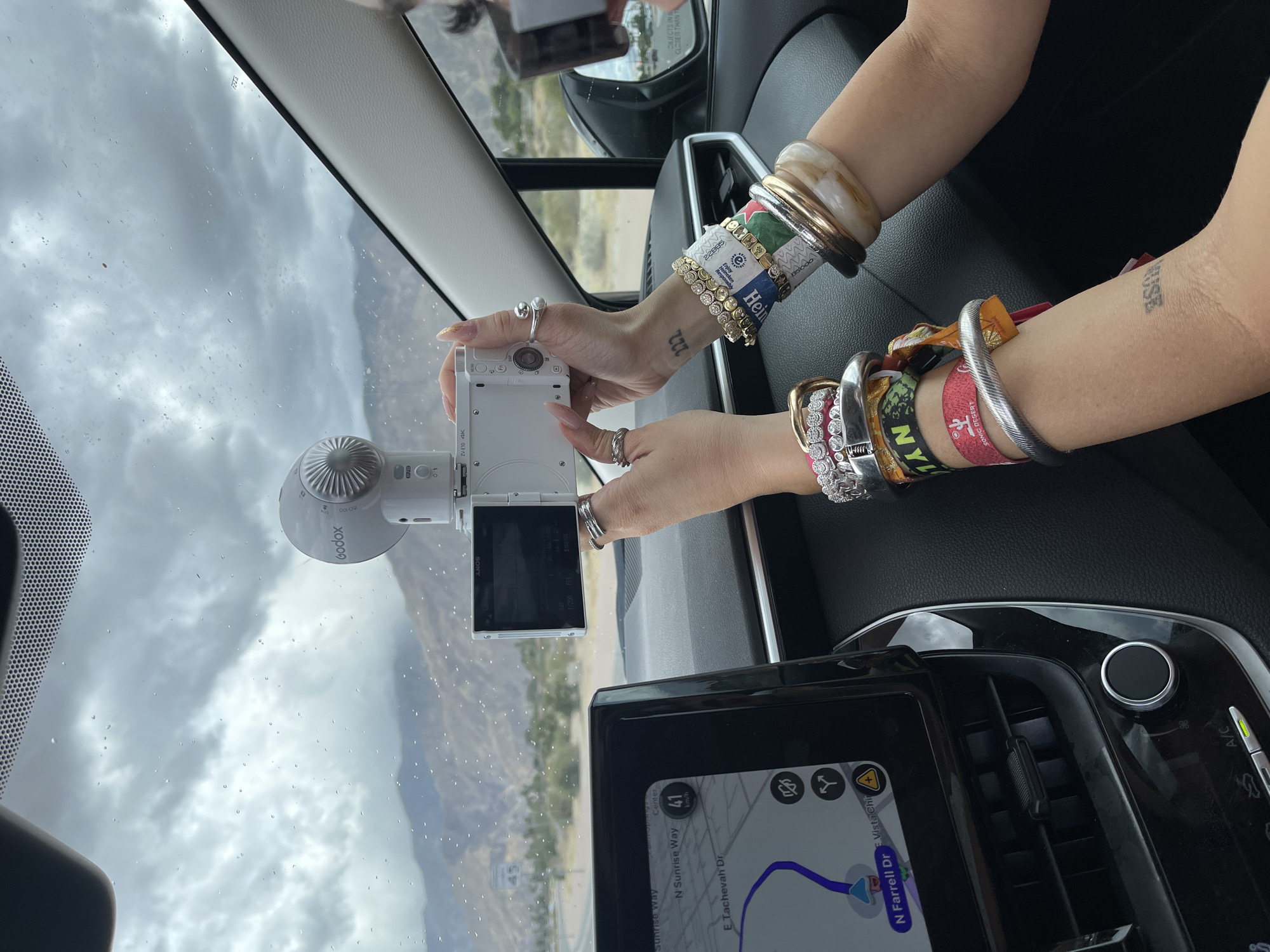 Here is a concise description of the image:
The photograph appears to be taken from the interior of a car, with a focus on the dashboard and windshield. A person's arms are visible, adorned with multiple bracelets and rings, holding a camera with its screen facing upwards. The camera is attached to a Godox device. A navigation screen displays a map indicating a route on North Sunrise Way towards North Farrell Drive in Palm Springs. The road is marked with a speed of 41 km/h. The environment outside the windshield suggests an outdoor setting, with overcast skies and mountainous terrain visible in the background. Tattoos are seen on the wrists. The overall setting suggests the person is capturing footage while driving.