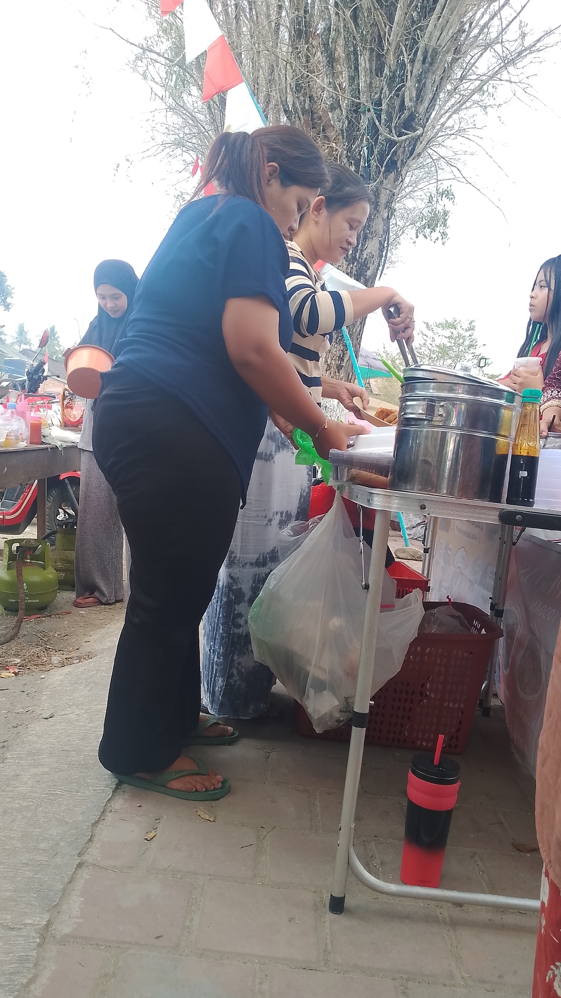 In the city of Karanglangu, Indonesia, a street scene unfolds. Three women are gathered around a food stall. The woman in the foreground, wearing a dark blue shirt and black pants, is turned away from the camera, her back to the viewer. She is wearing green flip-flops. To her right, another woman in a striped shirt is actively serving food from a large metal steaming pot. A third woman, wearing a headscarf and a pink shirt, stands further to the right, holding a drink. The food stall itself is set up on a metal table, with various items displayed, including a bottle of sauce and a plastic bag filled with goods. A green gas cylinder is visible on the ground to the left of the stall, and a red motorcycle is parked nearby. The background features a large tree with bare branches, possibly indicating a dry season, and flags are visible, one red and white, suggesting a national celebration. The ground is paved with light-colored tiles.