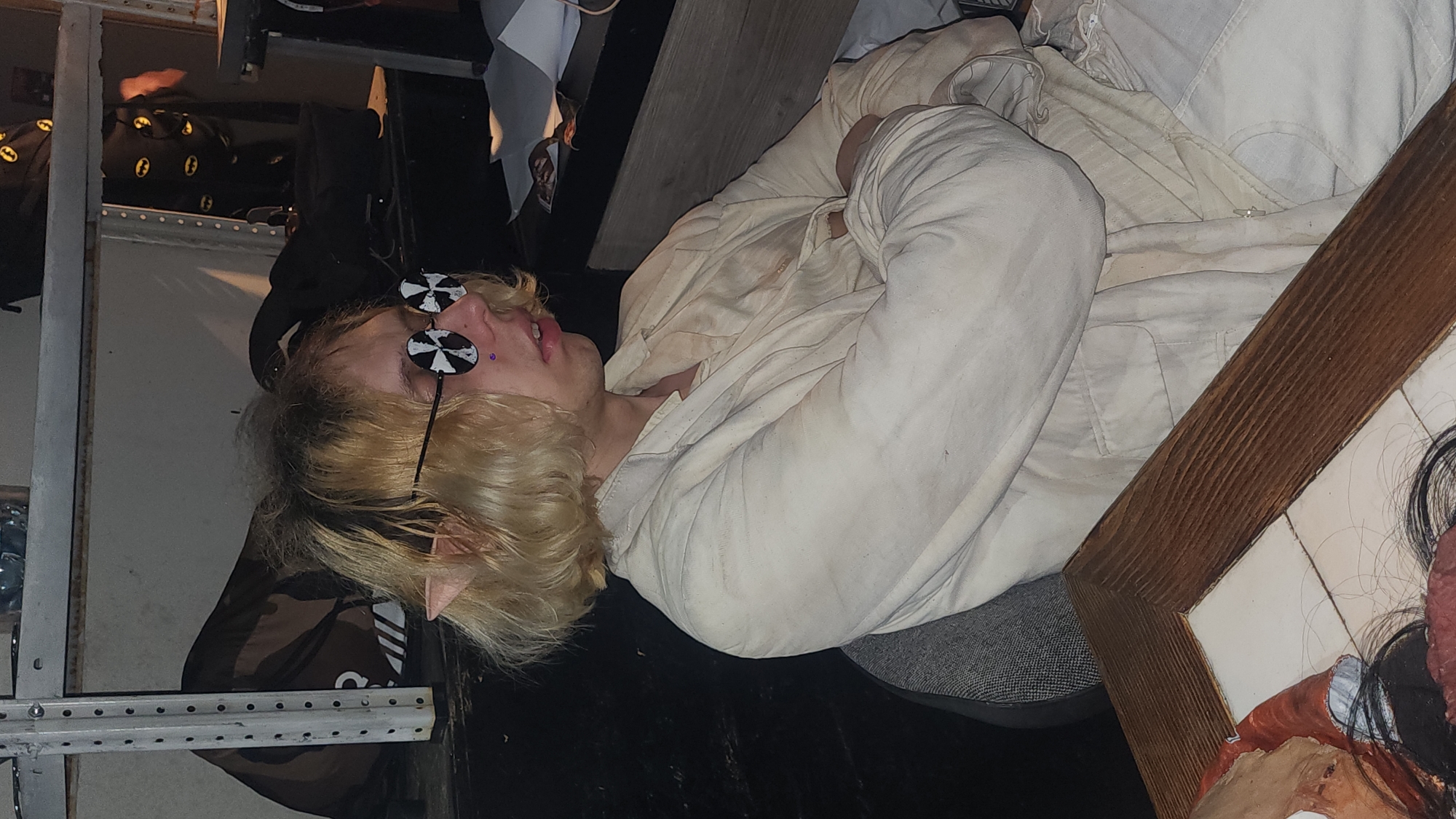 A person with blonde hair and pointed ears, wearing round black and white glasses and a light-colored jacket, is lying down in what appears to be an interior space. A piercing is visible near their lip. The environment seems cluttered, with various objects and structures visible in the background. The location in the city of Moscow, Russia, is not verifiable based on the visual data.