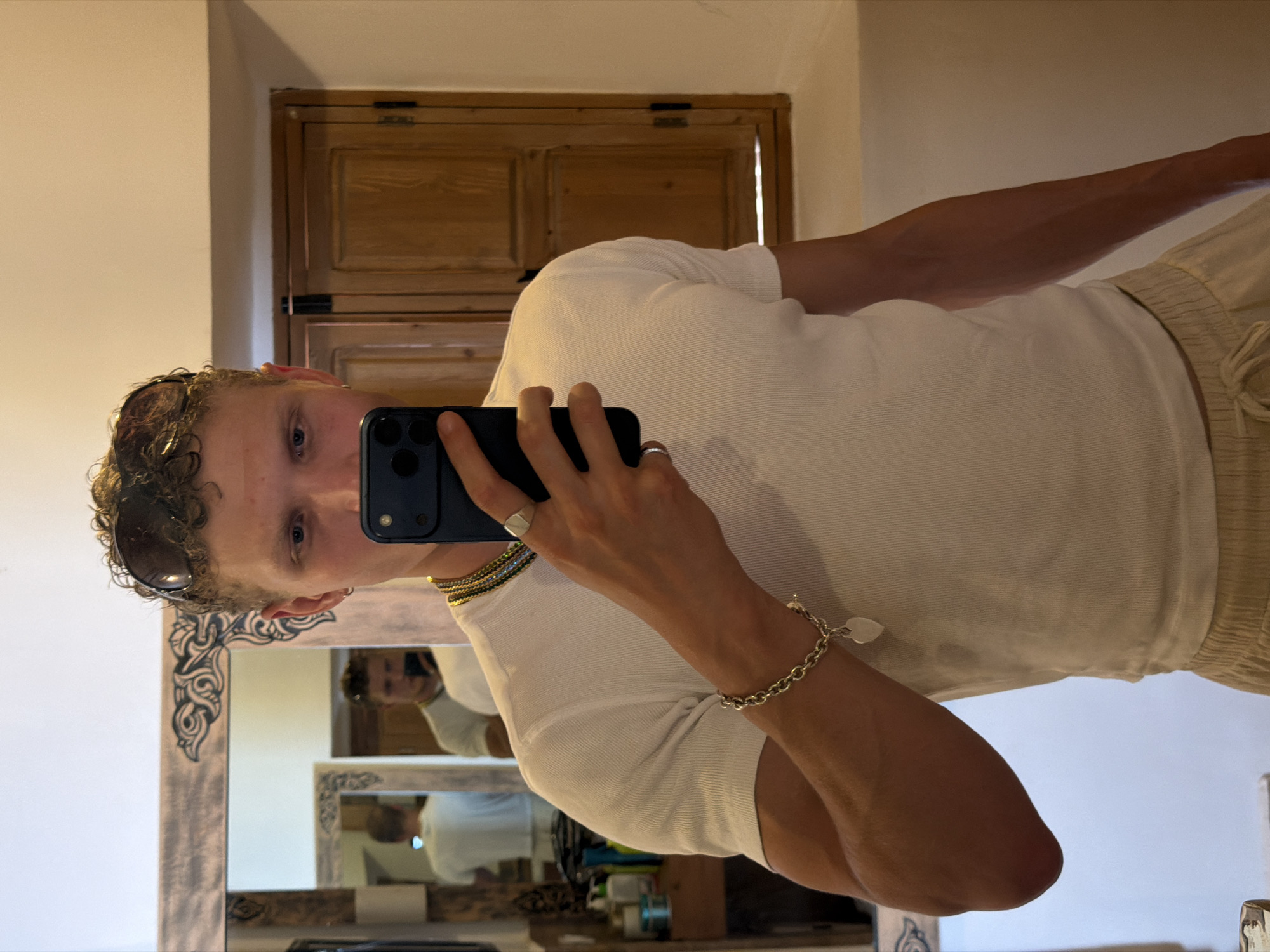 A young man with curly blonde hair and blue eyes takes a selfie in a mirror. He is wearing a white ribbed t-shirt, light-colored drawstring shorts, sunglasses perched on his head, a silver ring on his right index finger, a multi-colored beaded necklace, and a silver bracelet with a heart charm on his left wrist. Reflected in the mirror are two other men, one in a white t-shirt and the other also in a white t-shirt, taking a selfie. The environment appears to be a room with warm lighting, possibly a hotel bathroom or bedroom, with wooden cabinetry and a decorative mirror frame. The overall impression is casual and posed, indicative of a personal moment captured for social media.