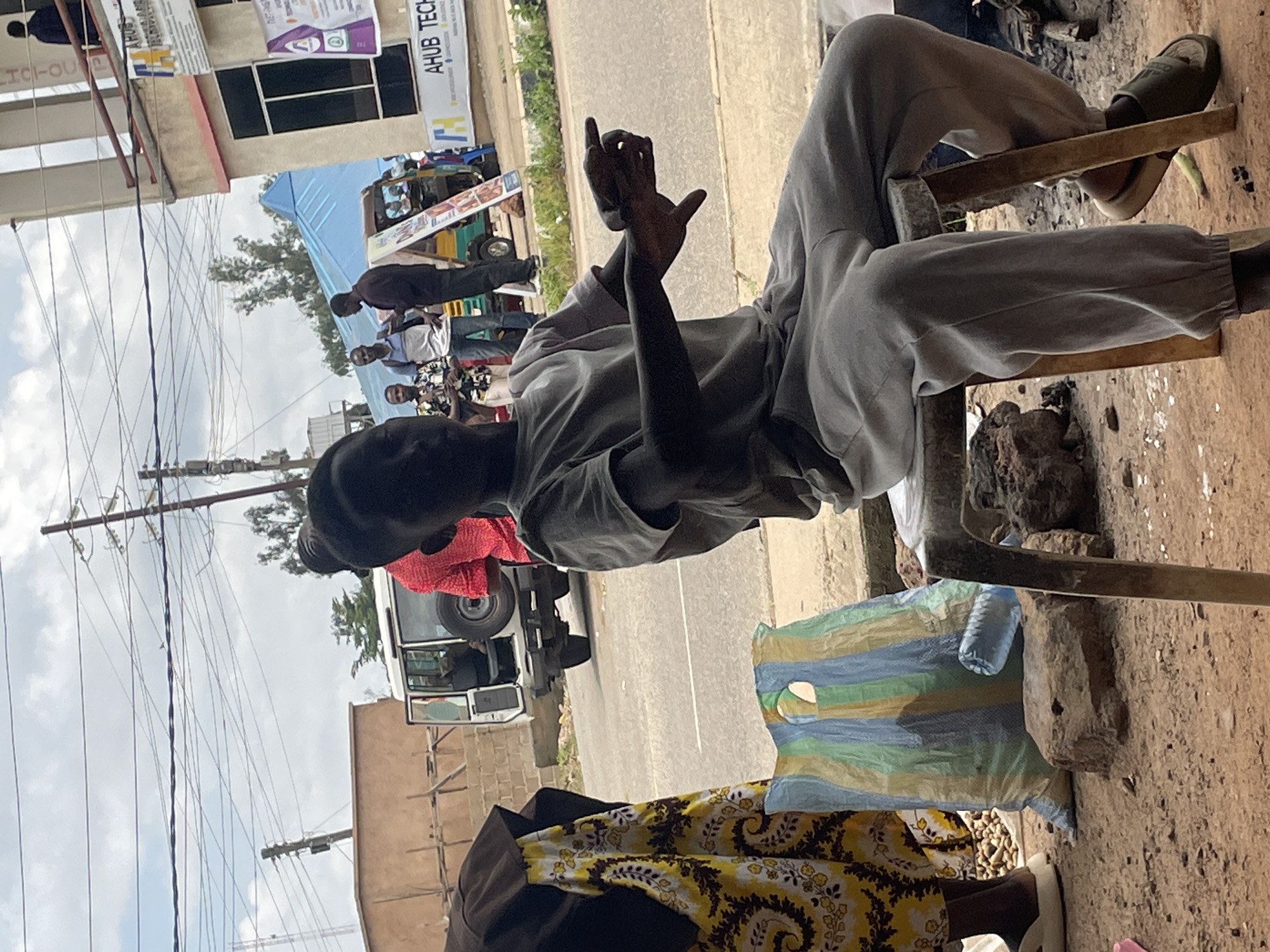 The image depicts a scene in Jos, Nigeria, with a young boy sitting on a stool in the foreground. He is wearing a light gray t-shirt and matching sweatpants, with a distinctive bun hairstyle. His arms are outstretched, and his right hand is pointing towards the right side of the frame. Behind him, on the left, a woman stands wearing a black headscarf and a yellow and black patterned dress. To the left of the woman is a large, multicolored striped bag. A white vehicle with its rear door open is parked on a paved road in the middle ground. Further in the background, several men are visible, some standing near a tricycle-like vehicle. One man, wearing a blue and white striped shirt, holds what appears to be a smartphone. The environment suggests an outdoor setting, possibly a roadside market or a busy street, with a building and overhead power lines visible in the background under a cloudy sky.