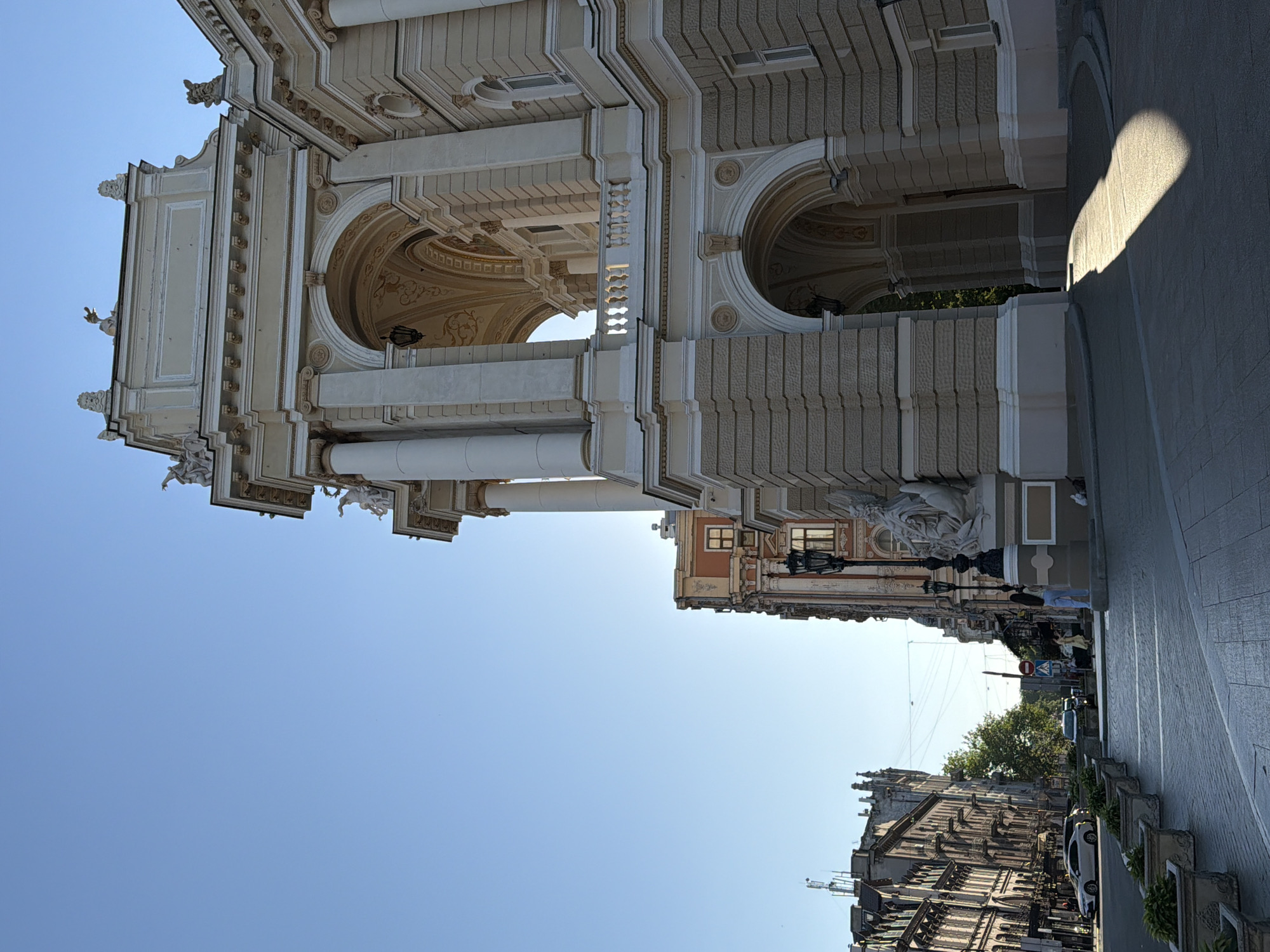 A low-angle, full shot captures the ornate facade of an opera house and a street scene in the city of Unknown, Unknown. The opera house, bathed in sunlight, features classical architecture with large arches, columns, and decorative sculptures. To the left, a street lined with several buildings, some with elaborate facades, leads into the distance. Cars are parked along the street, and a few people are visible walking. Potted plants are arranged along the sidewalk. The sky is clear and blue.
