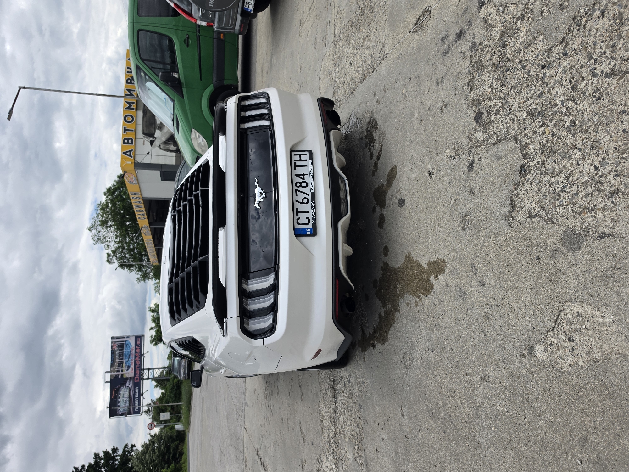 In the city of Tarnicheni, Bulgaria, a white Ford Mustang is parked on a weathered concrete surface, possibly a parking lot or area outside a car wash. The car is viewed from the rear, showcasing its distinctive taillights, a black spoiler, and louvers on the rear window. The license plate is clearly visible: CT 6784 TH. In the background, a green van is parked, and a car wash building with a yellow facade and "CARWASH" and "АВТОМИВКА" signage is present. A large billboard displaying an advertisement for "PAVEL BANYA DIANAmer.eu" is also visible in the distance. The sky is overcast with clouds. The concrete ground shows signs of wear and tear, with patches of dampness and embedded gravel.