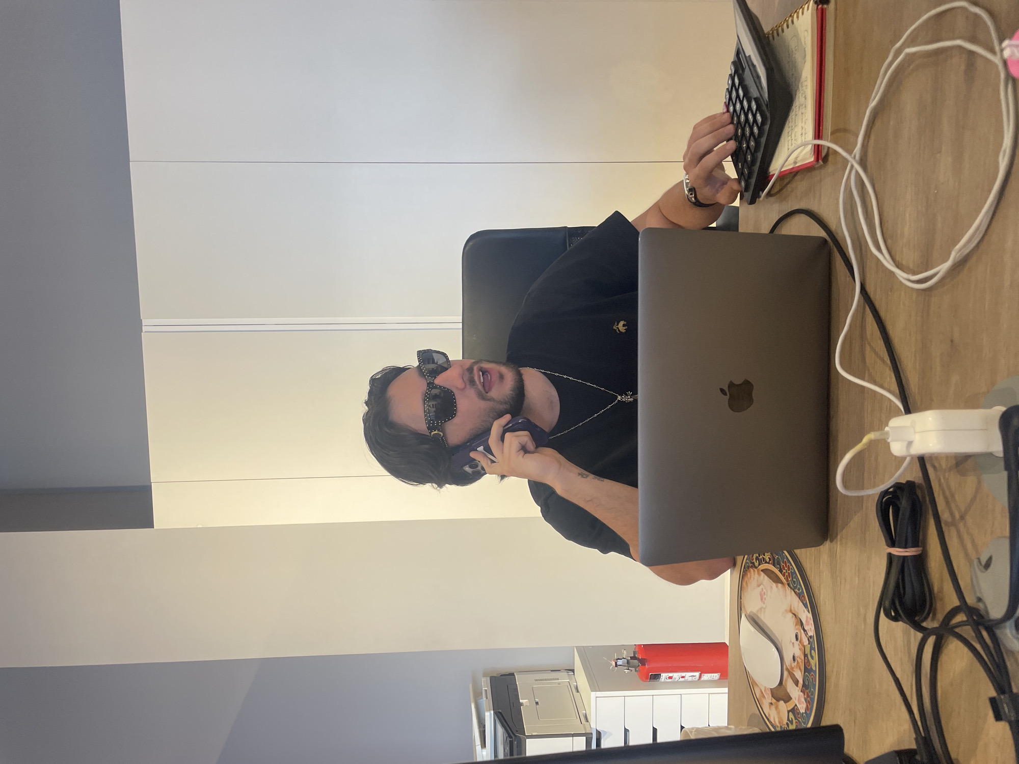 A person is sitting at a desk in an office setting, engaged in a phone call while simultaneously operating a calculator. The person is wearing sunglasses, a black t-shirt, and a chain necklace. A laptop is open in front of them on the desk. Other items on the desk include a notebook, cords, a charging block, and a decorative plate with an image of cats. A fire extinguisher and a printer are visible in the background. The location is within Mexico City, Mexico.