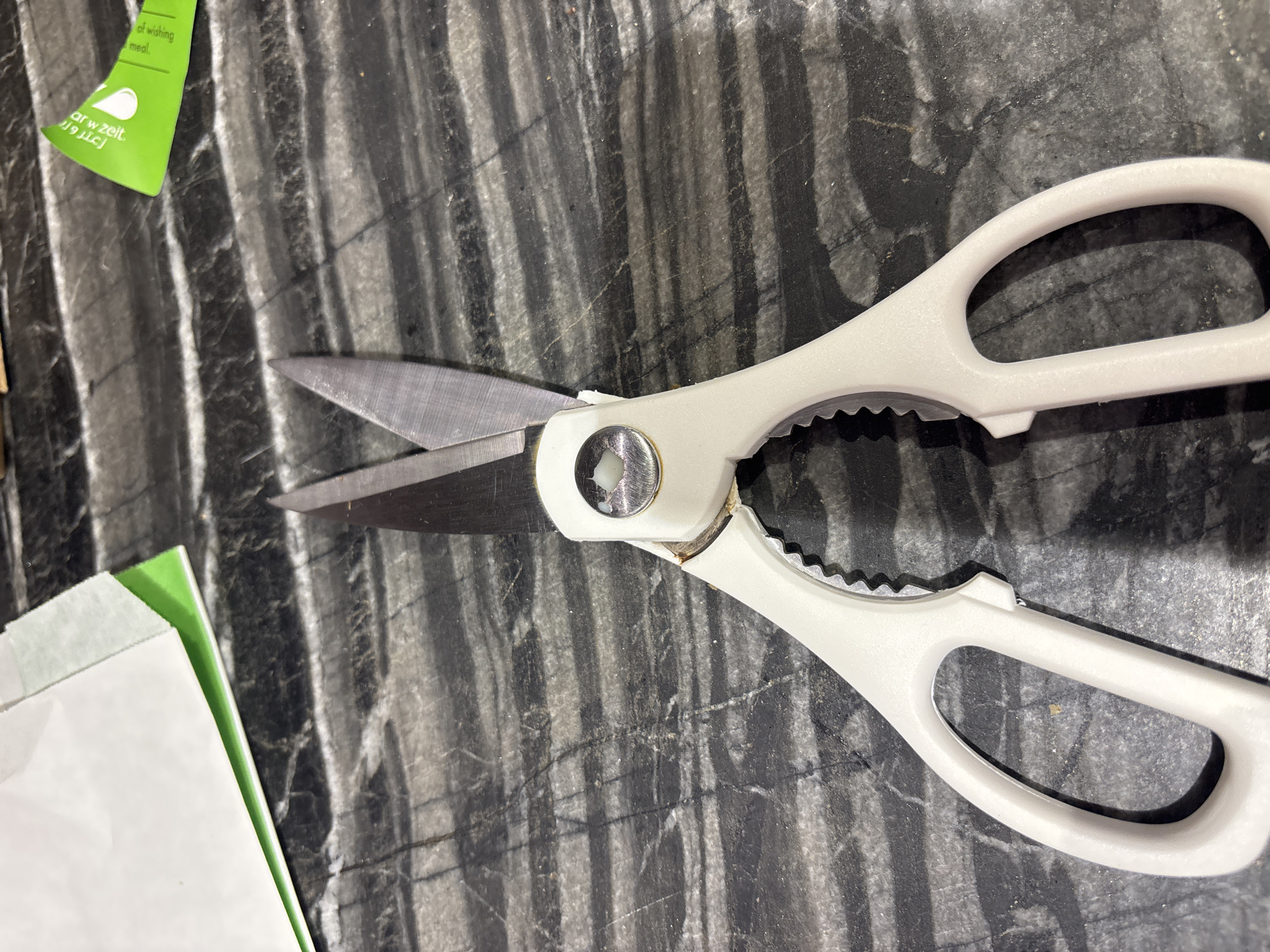 A pair of white-handled kitchen shears rests open on a dark, veined countertop. The shears are positioned in the center of the frame, with the blades pointing upwards and slightly towards the left. The handles are made of a matte white material, and the blades appear to be made of stainless steel. The countertop has a marbled pattern of black and grey, with white veining. In the upper left corner, a portion of a white envelope with a green edge is visible. In the upper right corner, a small, curved piece of green packaging with text in white is also present. The scene is indoors, and the lighting suggests it is a well-lit environment. There is no indication of the location being Dubai, United Arab Emirates, or any specific city.