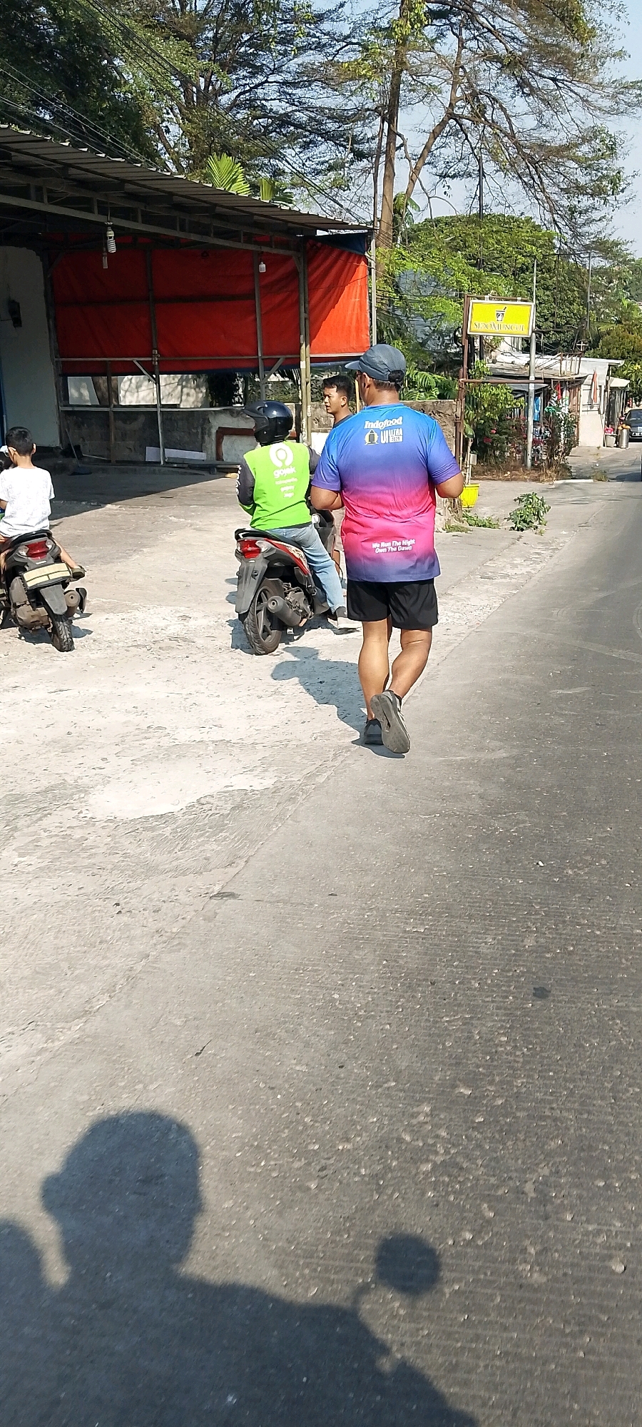 A man wearing a blue and pink gradient t-shirt and black shorts is running on the side of a road. He has a gray baseball cap on. Behind him, a person in a bright green vest with "Gojek" branding is sitting on a scooter, wearing a black helmet. To the left, another person is sitting on a scooter, wearing a white t-shirt. A building with a red tarp covering its front is visible on the left side of the image. A yellow sign with Indonesian text is posted on a pole to the right. The ground is a mix of asphalt and concrete. The scene appears to be in Depok, Indonesia, as indicated by the presence of Gojek, a ride-sharing company popular in the region.