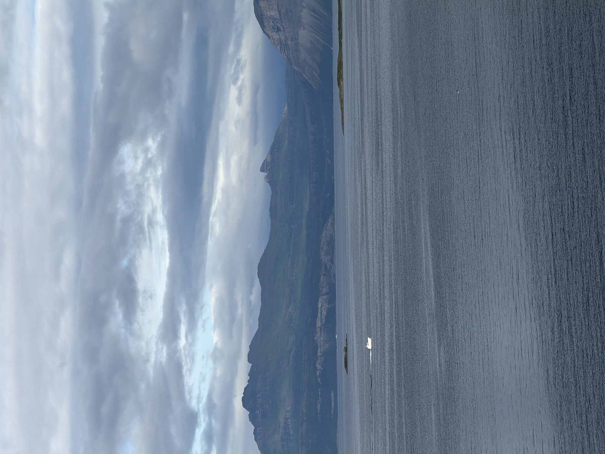 This is a wide shot of a calm, dark body of water with a mountainous coastline in the background, under a cloudy sky. The water is a deep blue-grey, with subtle ripples creating horizontal lines. In the middle distance, a small white boat is visible, creating a white wake as it moves from left to right. A small, tree-covered island with a few buildings is situated to the right of the boat. The coastline is dominated by large, green, and rocky mountains, with a flatter, layered rock formation visible in the foreground of the mountains. The sky is filled with dark, dramatic clouds, with a few breaks of lighter sky showing through. The scene is serene and natural. The location is believed to be the Vågsfjorden, near Harstad, Norway.