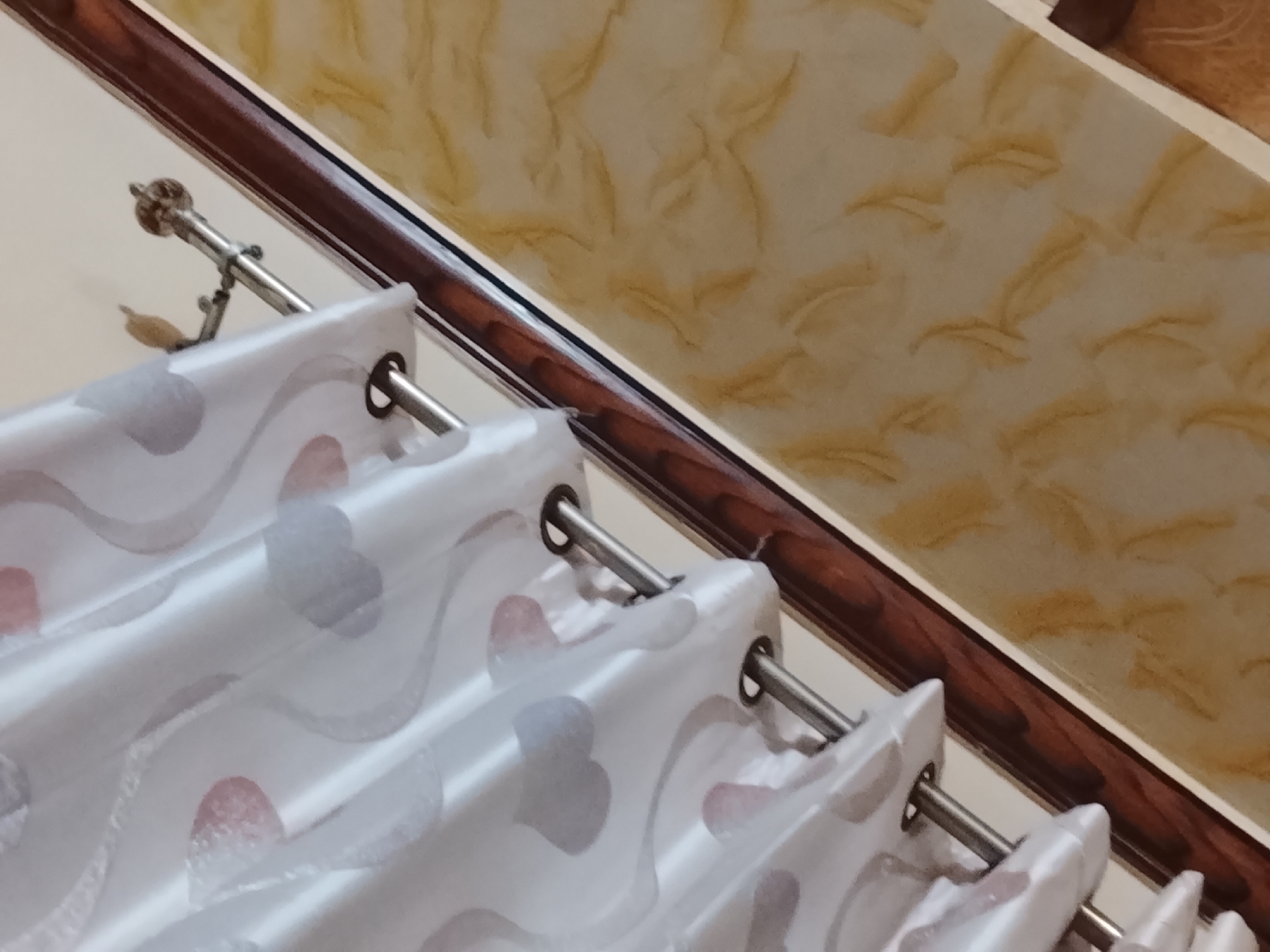 A low-angle close-up shot shows a curtain suspended from a rod, positioned below a wall. The curtain, featuring a white base with heart-shaped patterns in red and gray, hangs loosely from the rod via silver rings. The rod is also silver and has a decorative finial. Above the rod, a narrow, dark brown trim runs along the top of the wall. The wall itself is off-white and has a yellow and brown pattern resembling marbling or swirls. The image was taken indoors. The location in the city of Multan, Pakistan is not determinable from the image. There are no people visible.