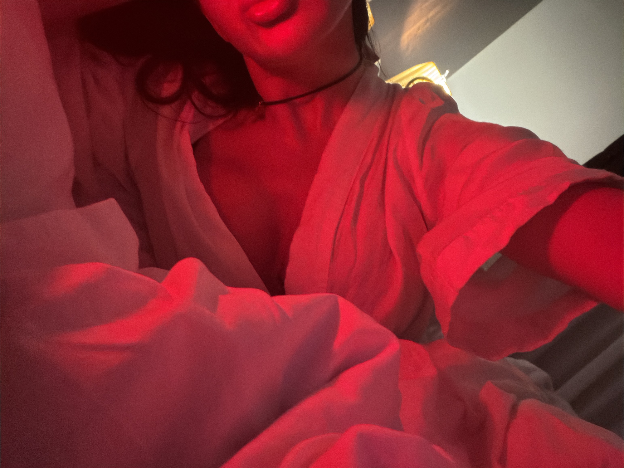 The image depicts a person in a white robe lying in bed. The lighting is heavily filtered with a red hue. The person is wearing a choker necklace. The background is out of focus but appears to be a room interior. The location is unknown; there is no verifiable connection to Boreti, Montenegro.