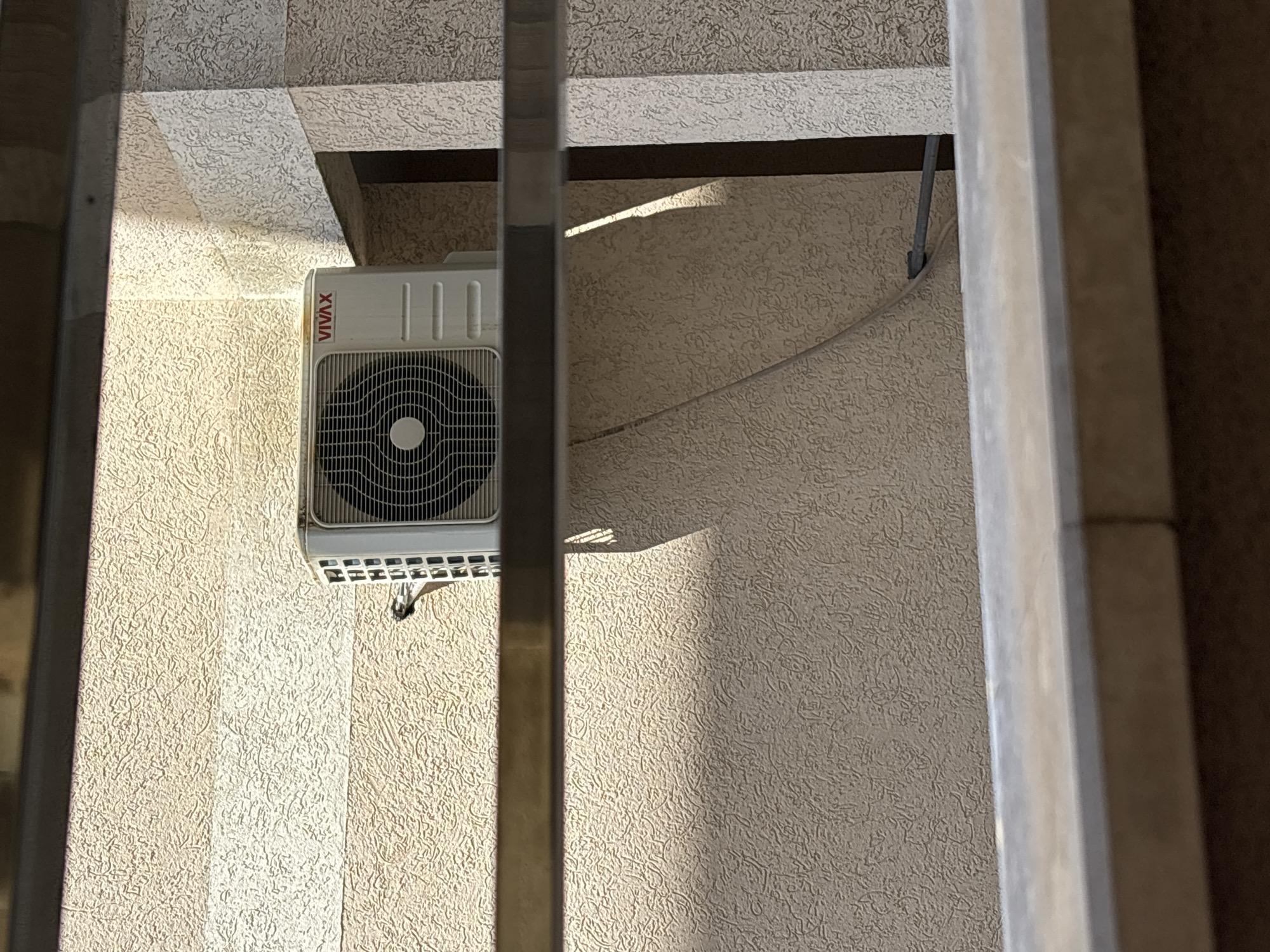 A close-up shot, likely taken in Budva, Montenegro, reveals a VIVAX air conditioning unit mounted on a textured, light-colored wall. The unit is partly obscured by a window frame. A hose or cable extends from the unit. The lighting indicates daylight conditions, with shadows cast on the wall. The view is constricted by the window frame, suggesting an indoor perspective.